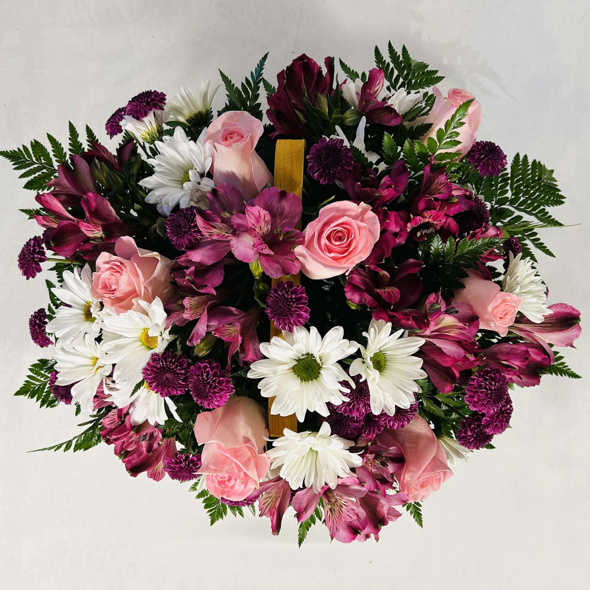 Round arrangement of pink roses, dark purple blooms, and white daisies in a basket, viewed from above