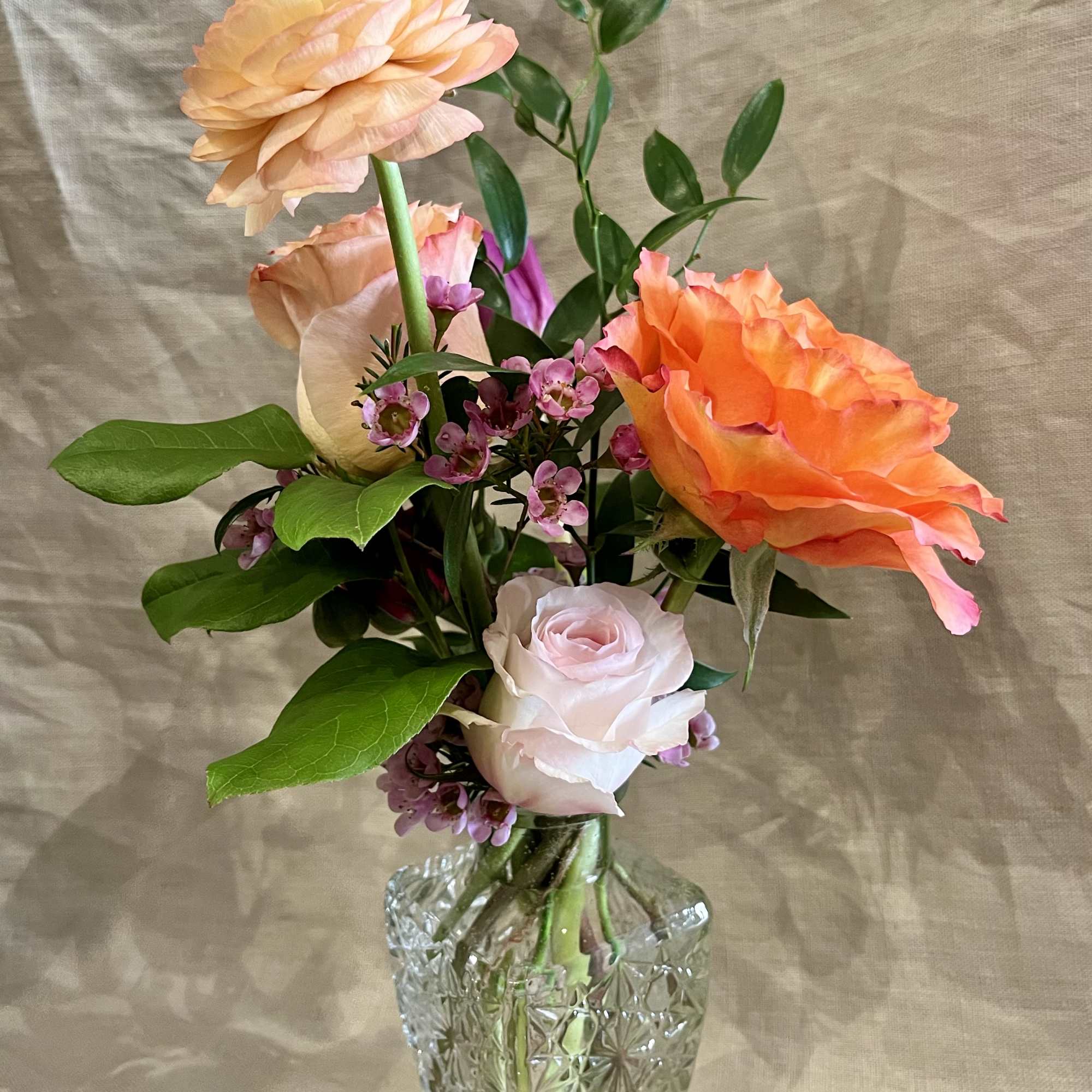 Small glass bud vase with peach ranunculus, orange and pale pink roses, and tiny purple flowers.