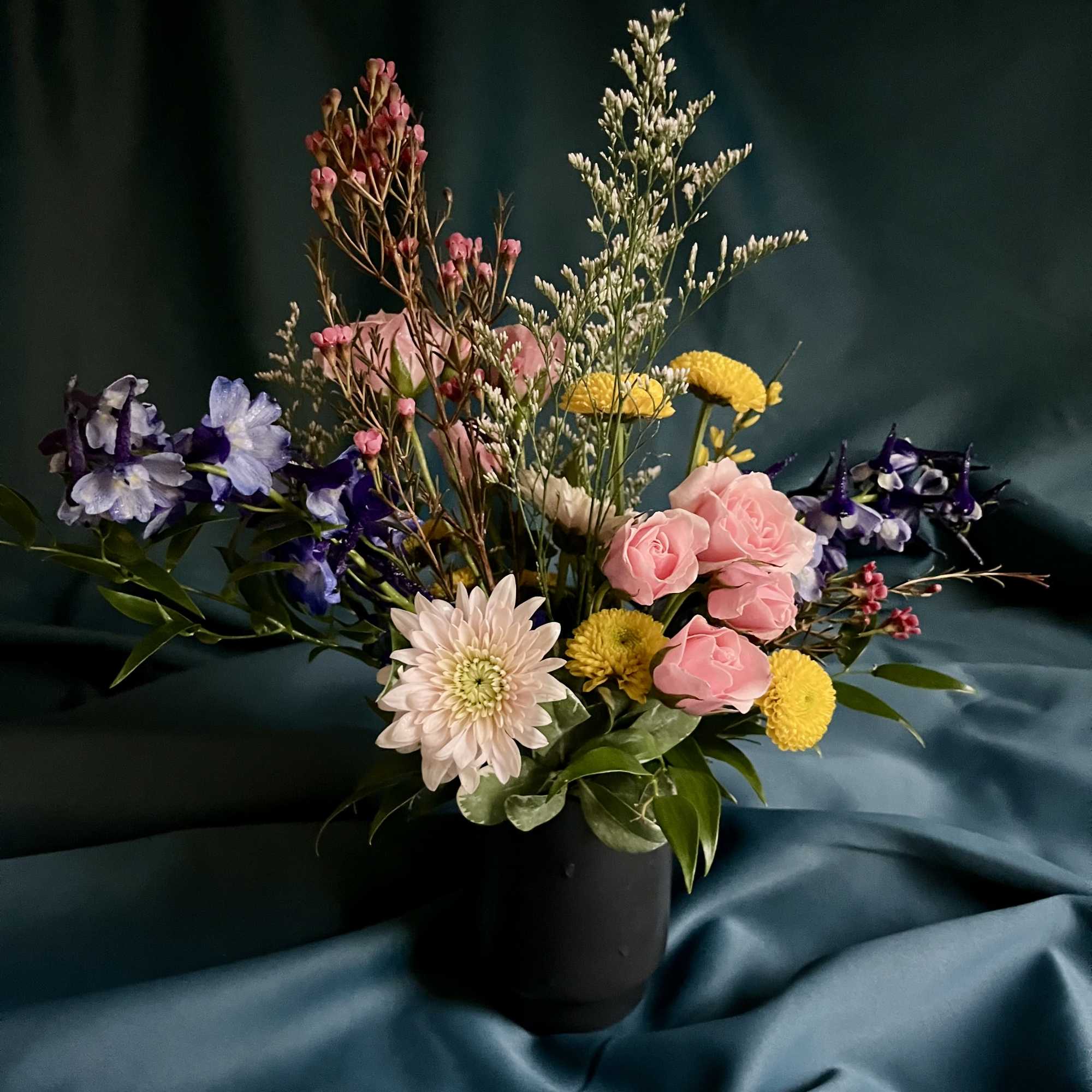 Mixed arrangement of pink roses, blue blooms, and yellow button flowers in a black vase on teal fabric