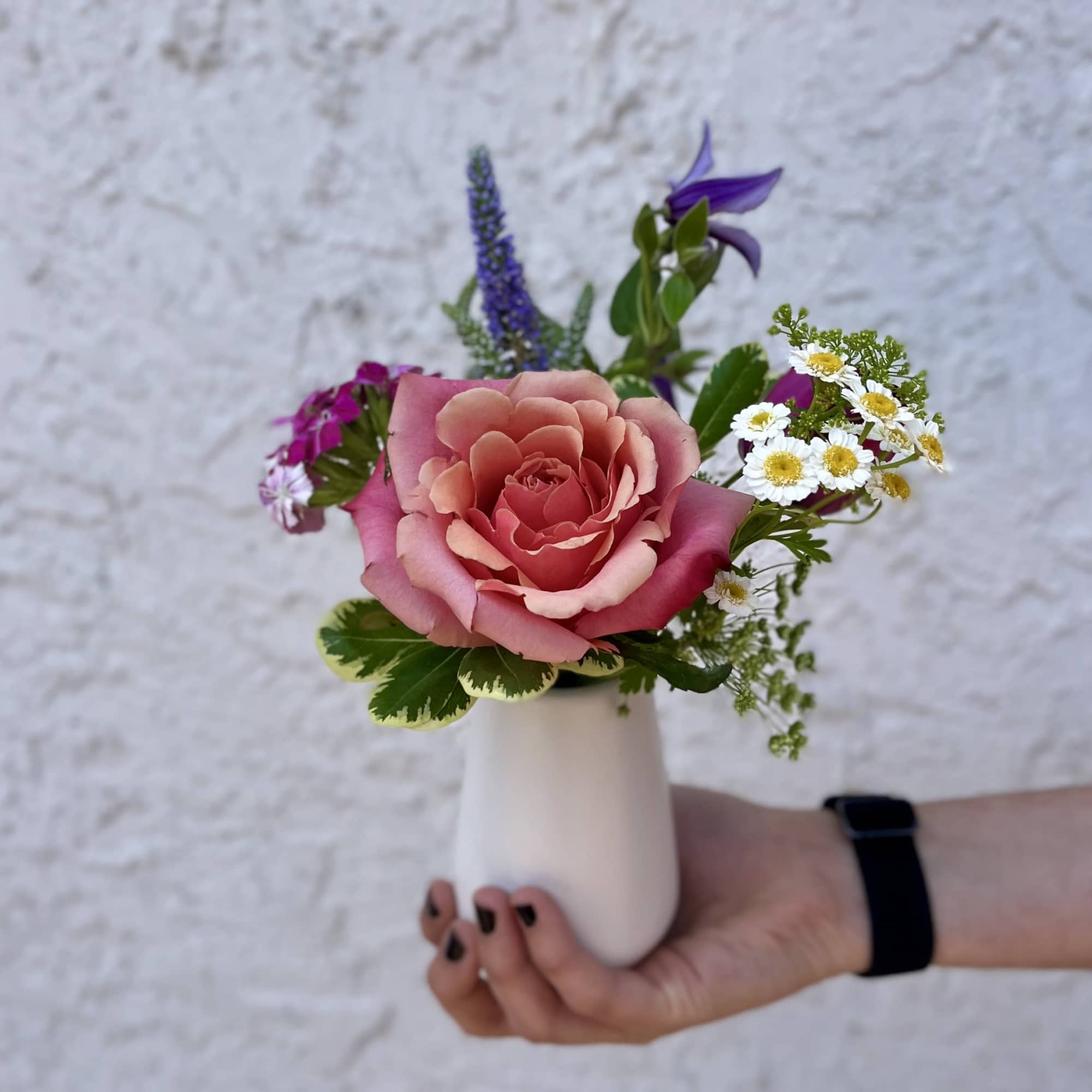 Small bud vase arrangement with a peach rose, white daisies, and purple blooms held in one hand