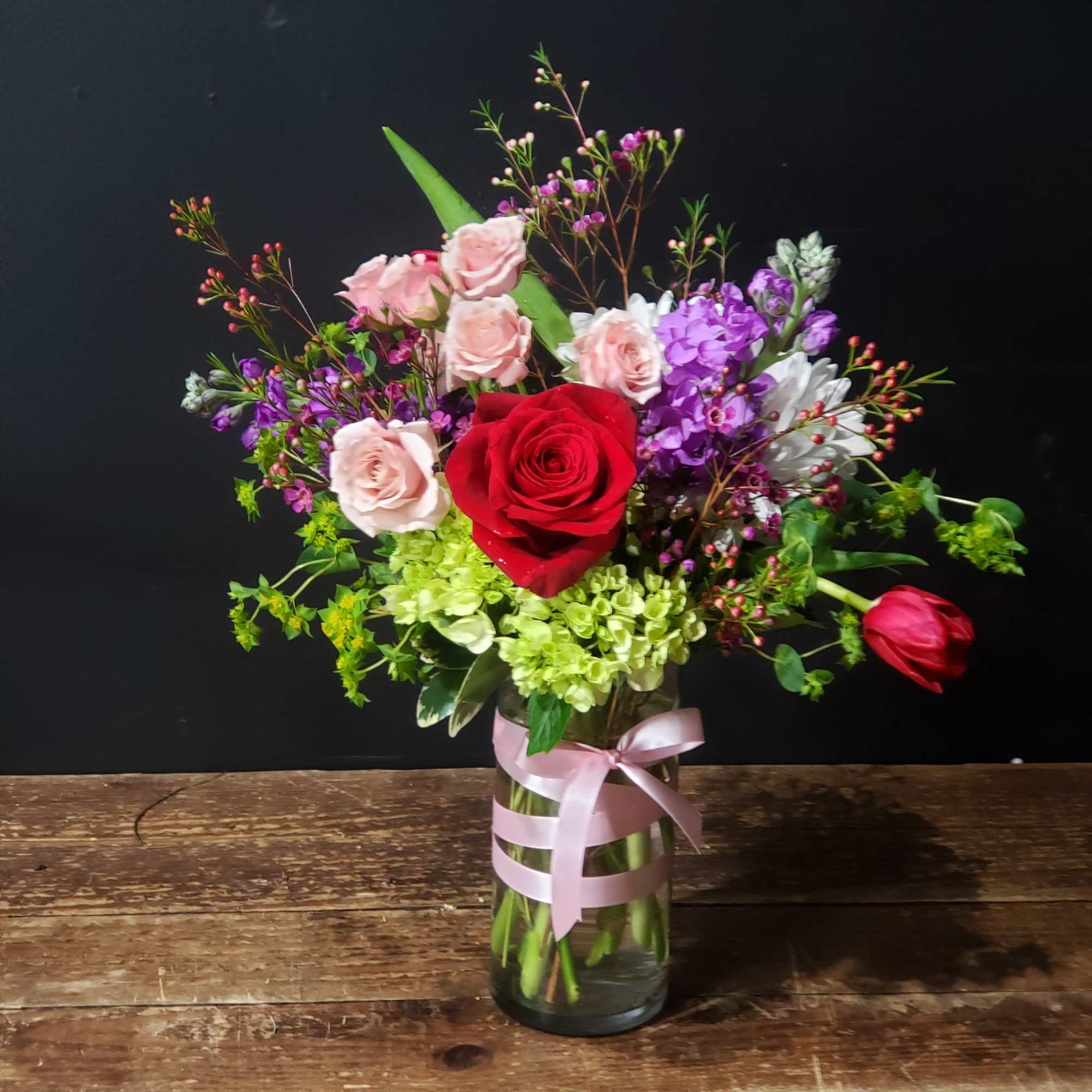 Mixed bouquet of roses and purple flowers in a glass vase with a pink ribbon