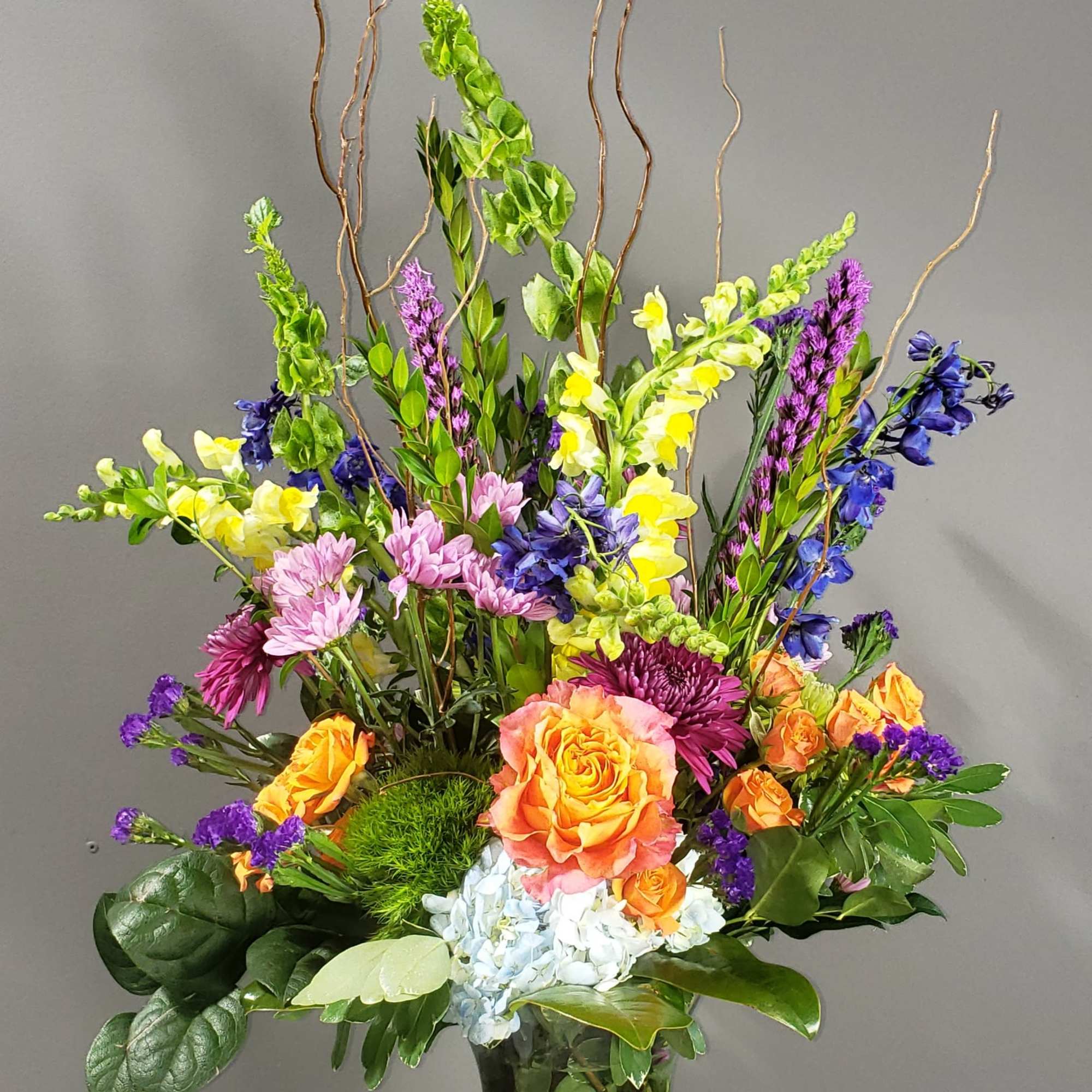 Tall mixed bouquet in a glass vase with orange, yellow, purple, and pink flowers