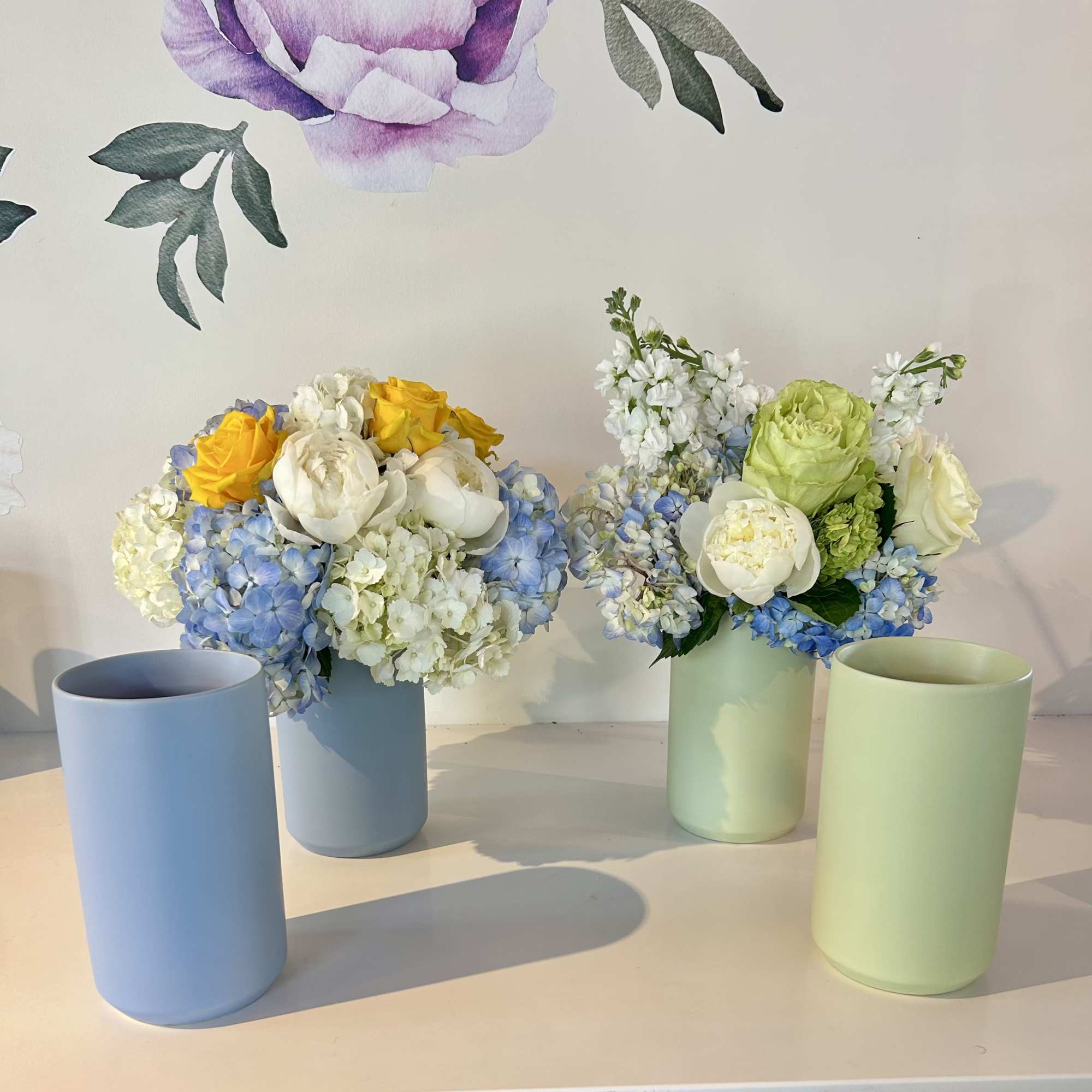 Two pastel floral arrangements in cylindrical vases with blue, white, yellow, and green blooms.