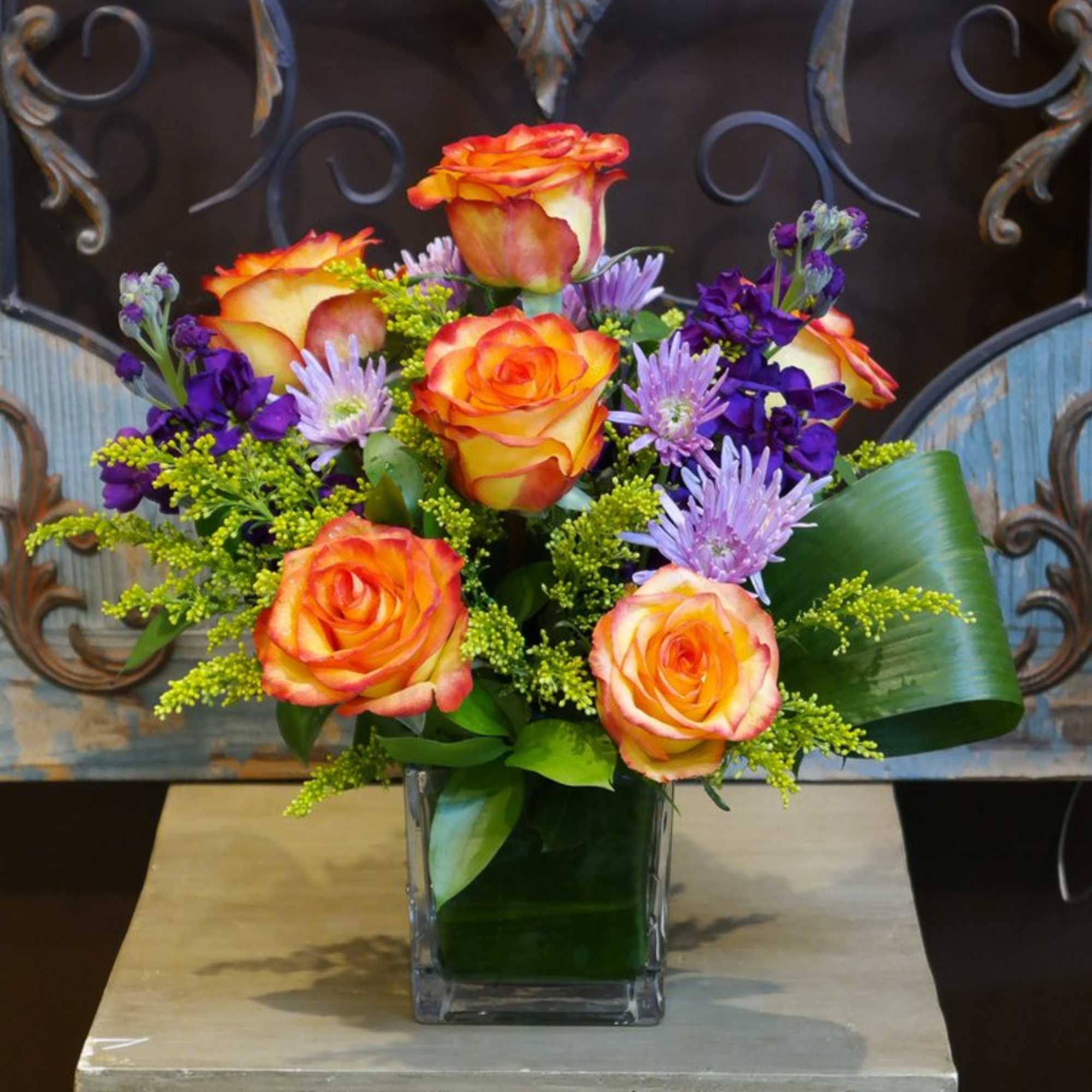 Bright arrangement of orange roses with purple and lavender flowers in a square glass vase
