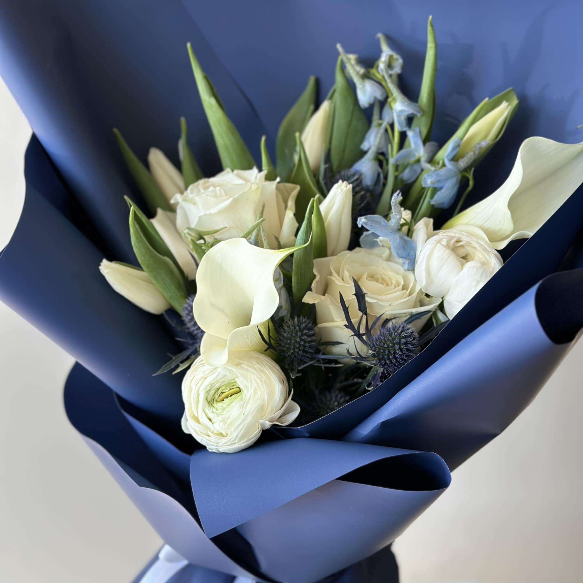Bouquet of white roses, calla lilies, and blue thistle wrapped in navy paper