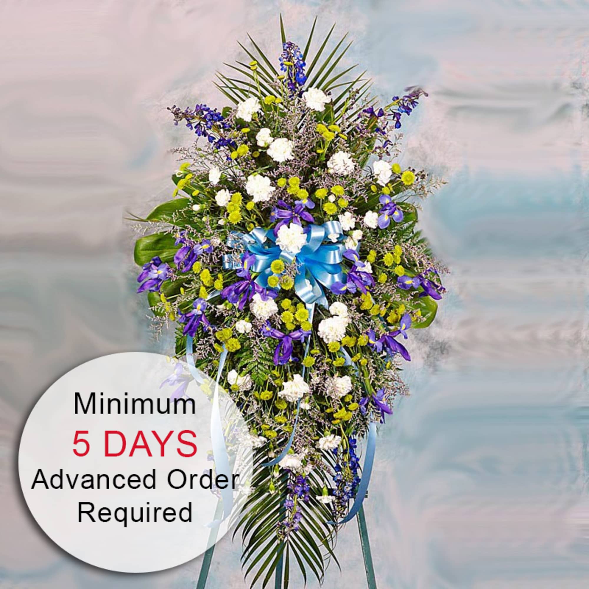 Tall standing spray with blue and purple flowers, white carnations, yellow mums, and blue bow on an easel.