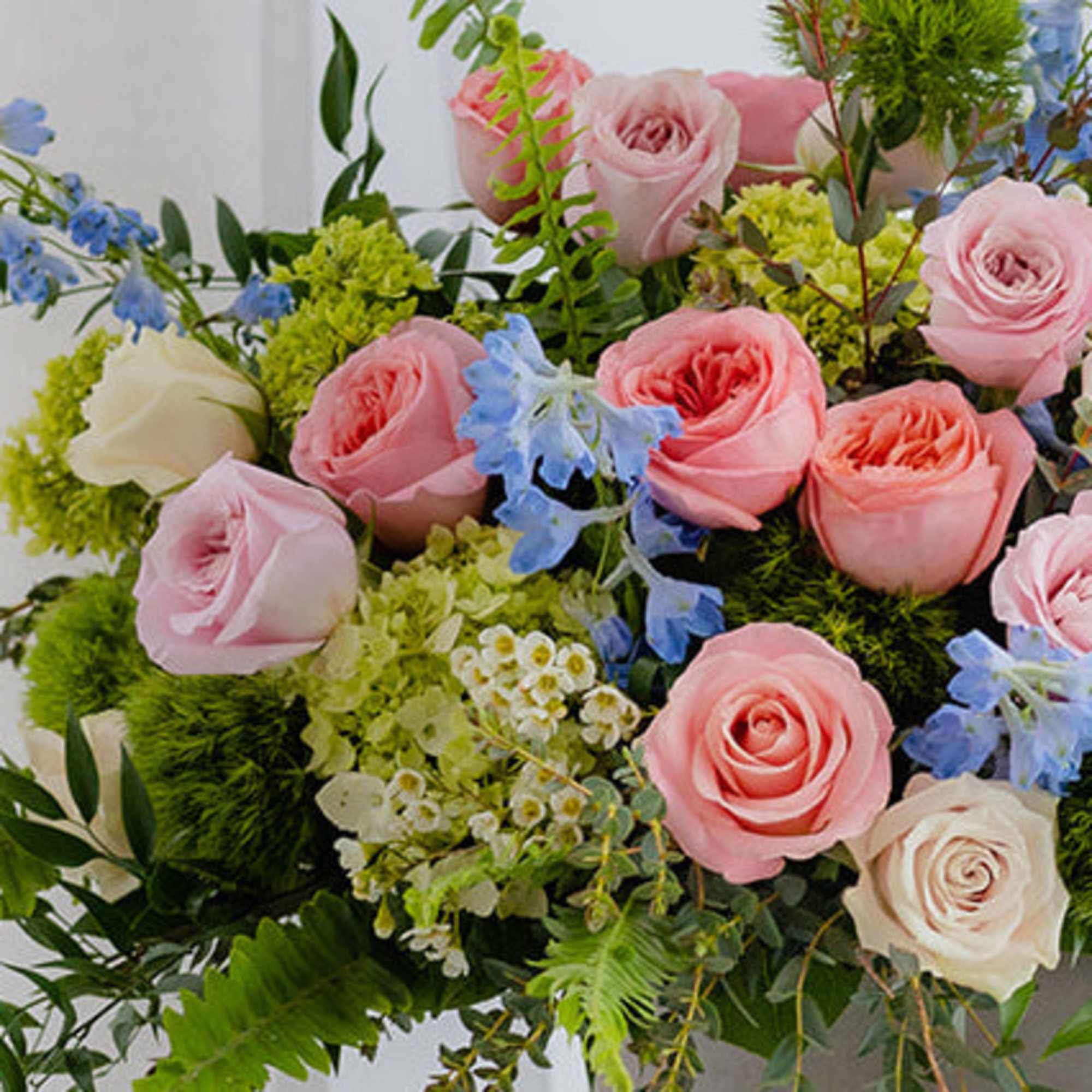 Pastel arrangement of pink and cream roses with blue flowers and green accents in a low container