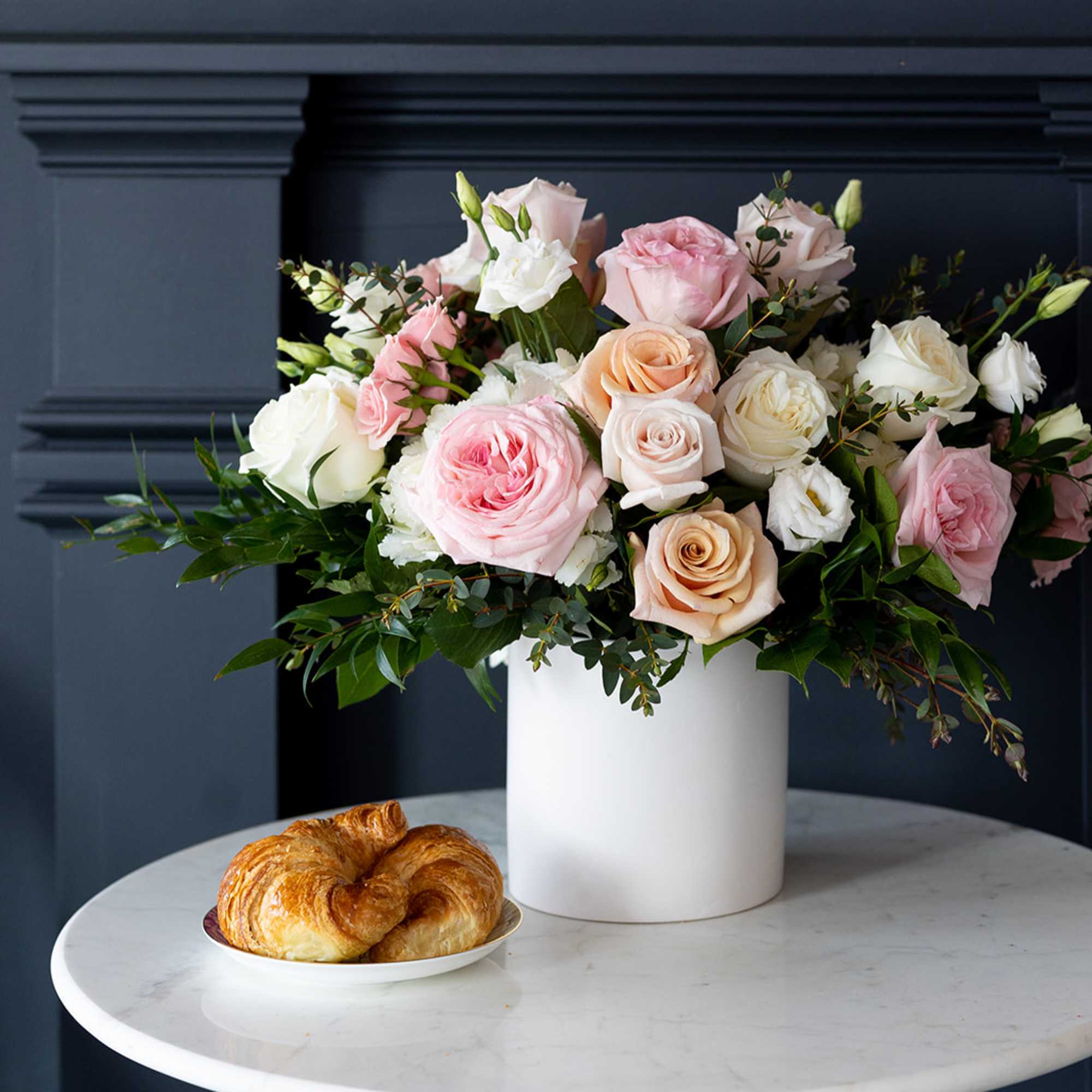 Pastel rose arrangement in a white cylindrical vase with a croissant on a plate