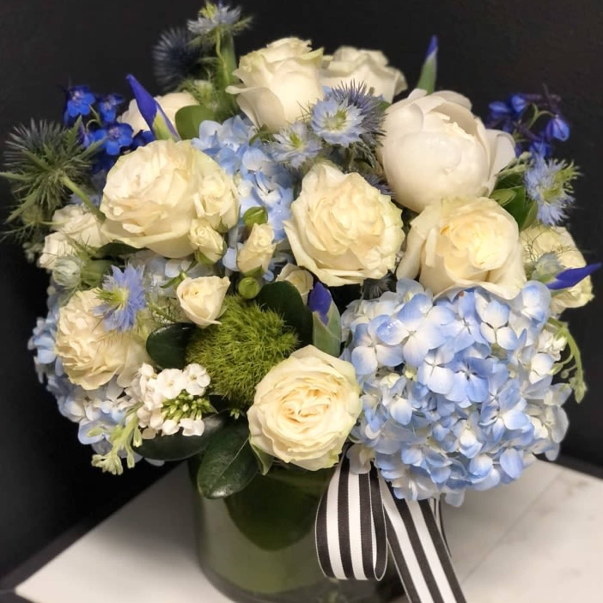 Cream roses and blue hydrangeas arranged in a glass vase with a striped ribbon.