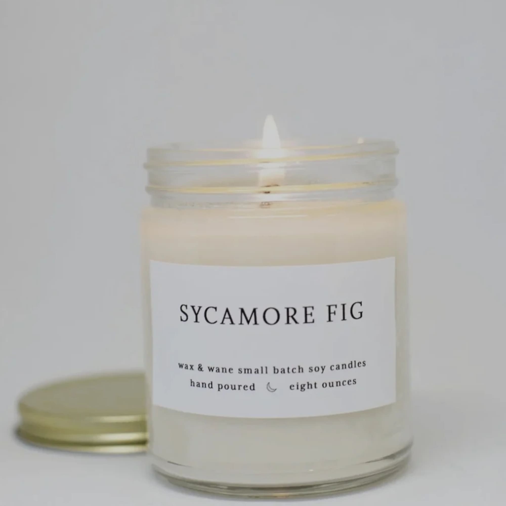 Lit white soy candle in a clear glass jar with Sycamore Fig label and gold lid beside it.