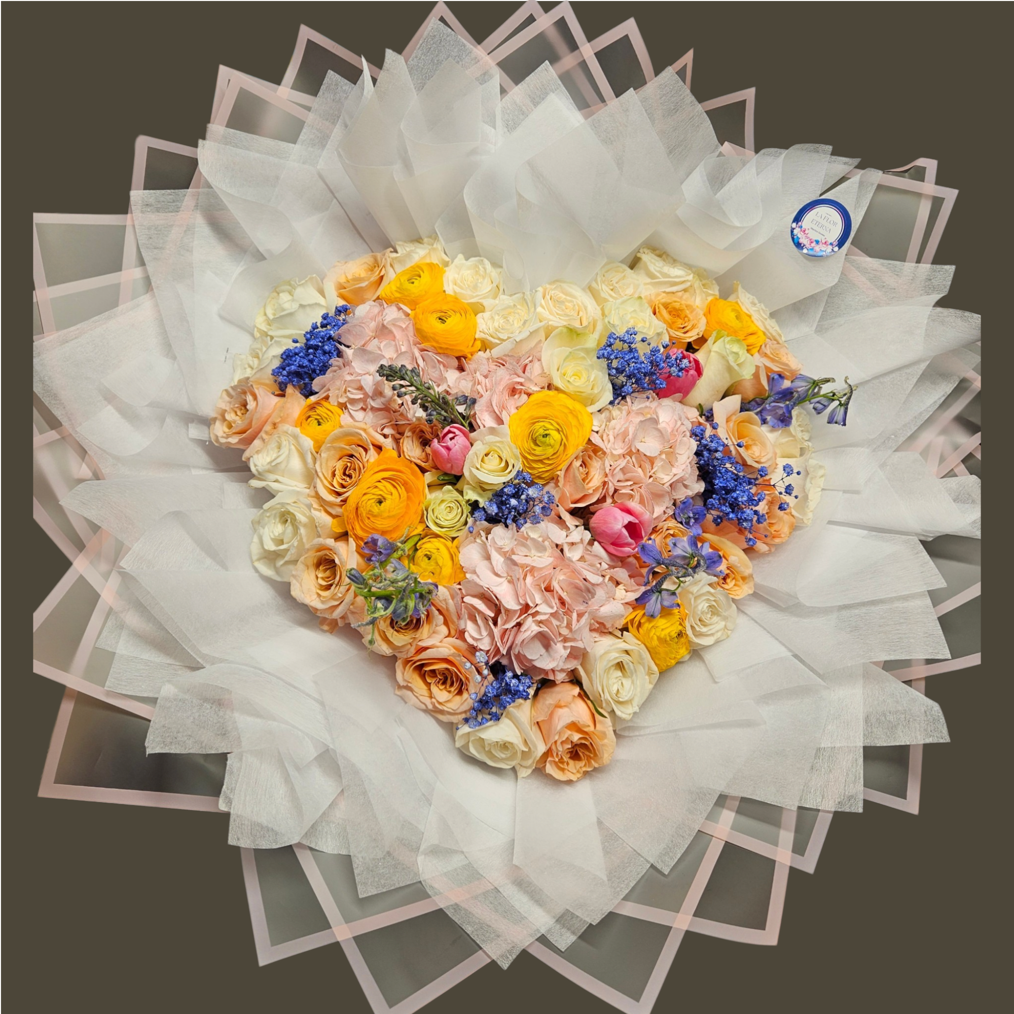 Heart-shaped bouquet of pastel roses, hydrangeas, ranunculus, and blue flowers wrapped in layered white paper.