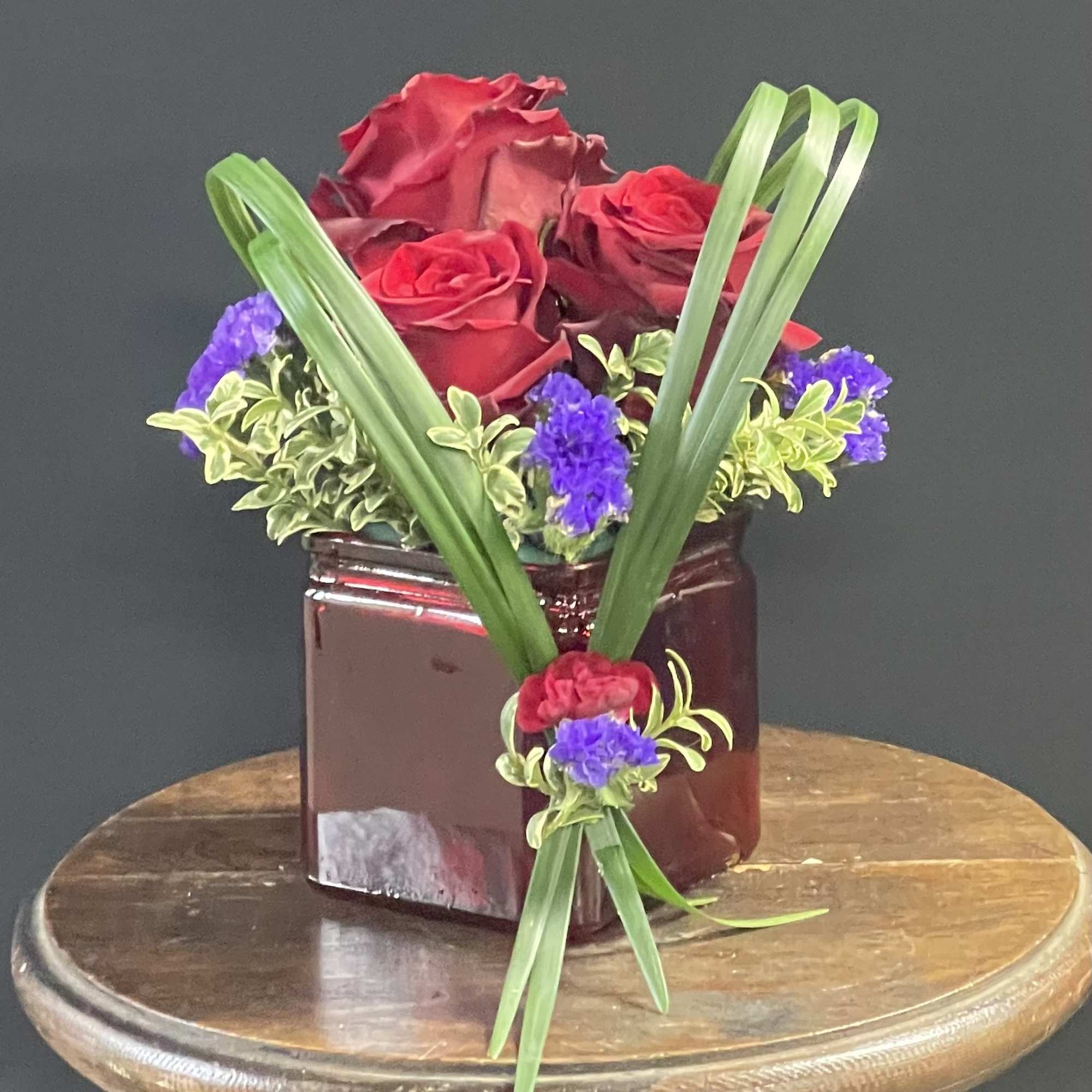 Red roses in a burgundy vase with purple accents and long green leaves