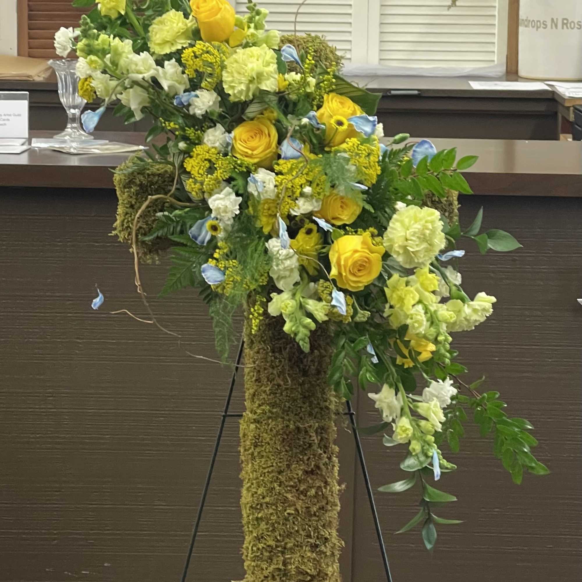 Tall floral arrangement with yellow roses, white blooms, and blue accents on a moss-covered stand.