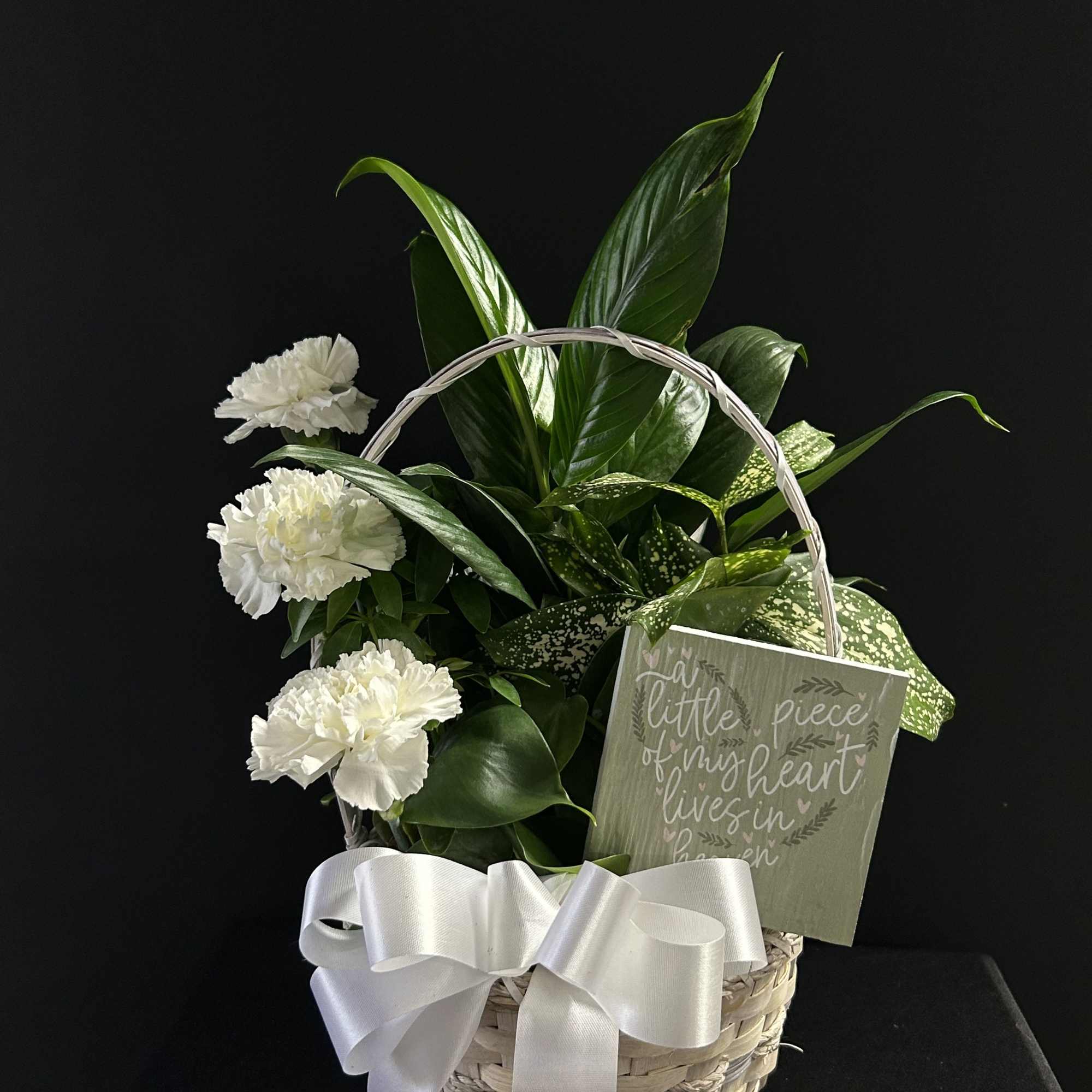 White carnations and green foliage in a wicker basket with a ribbon and sympathy card