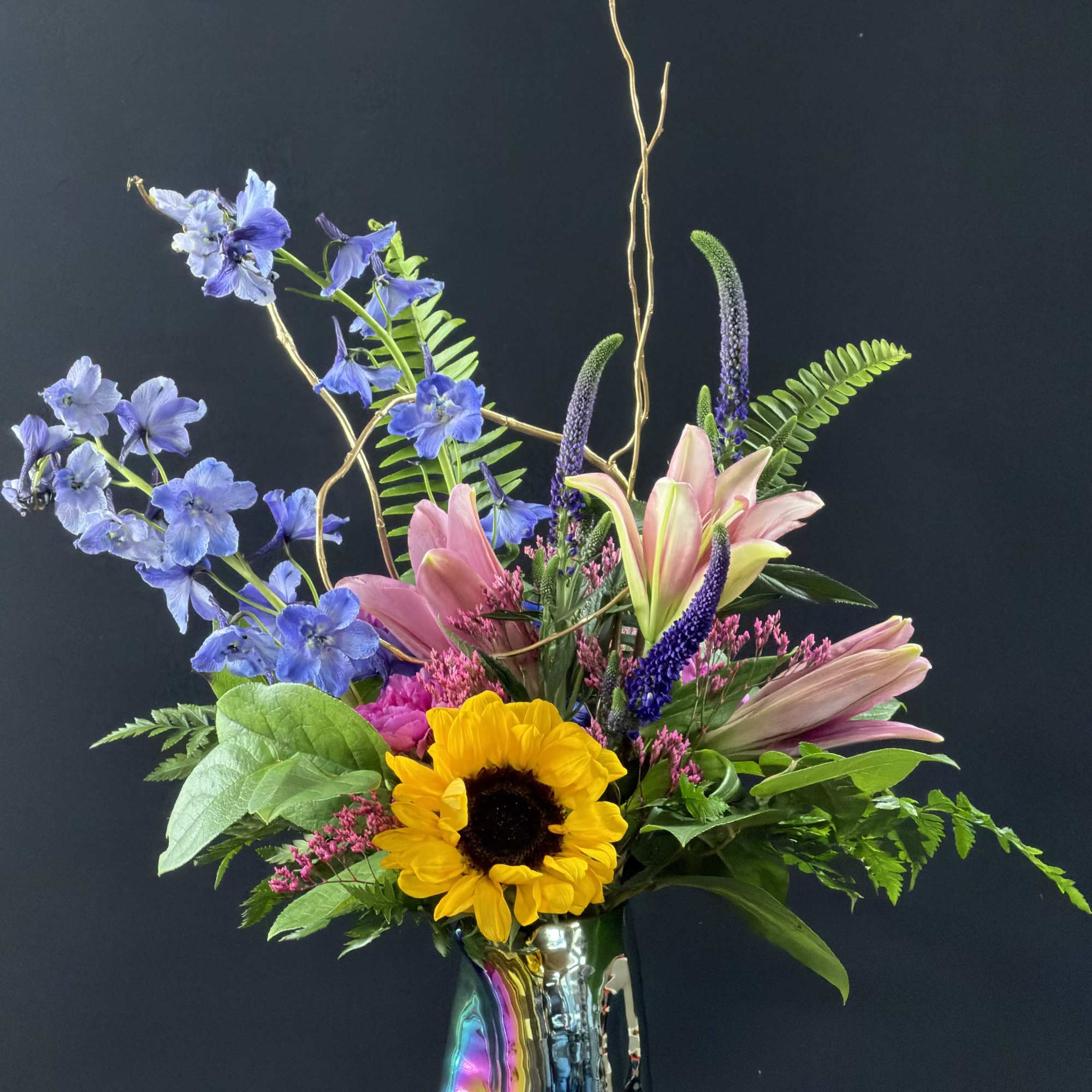 Tall mixed arrangement with pink lilies, blue delphinium, and a sunflower in an iridescent vase