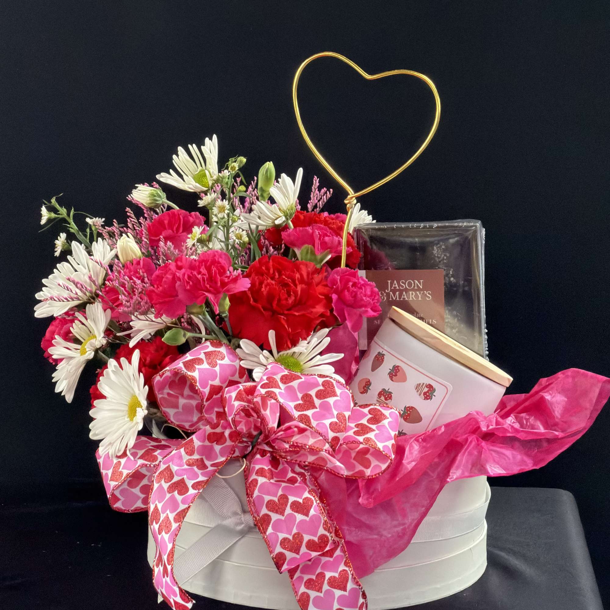 Pink and white flower arrangement with a heart topper and gift items in a round box