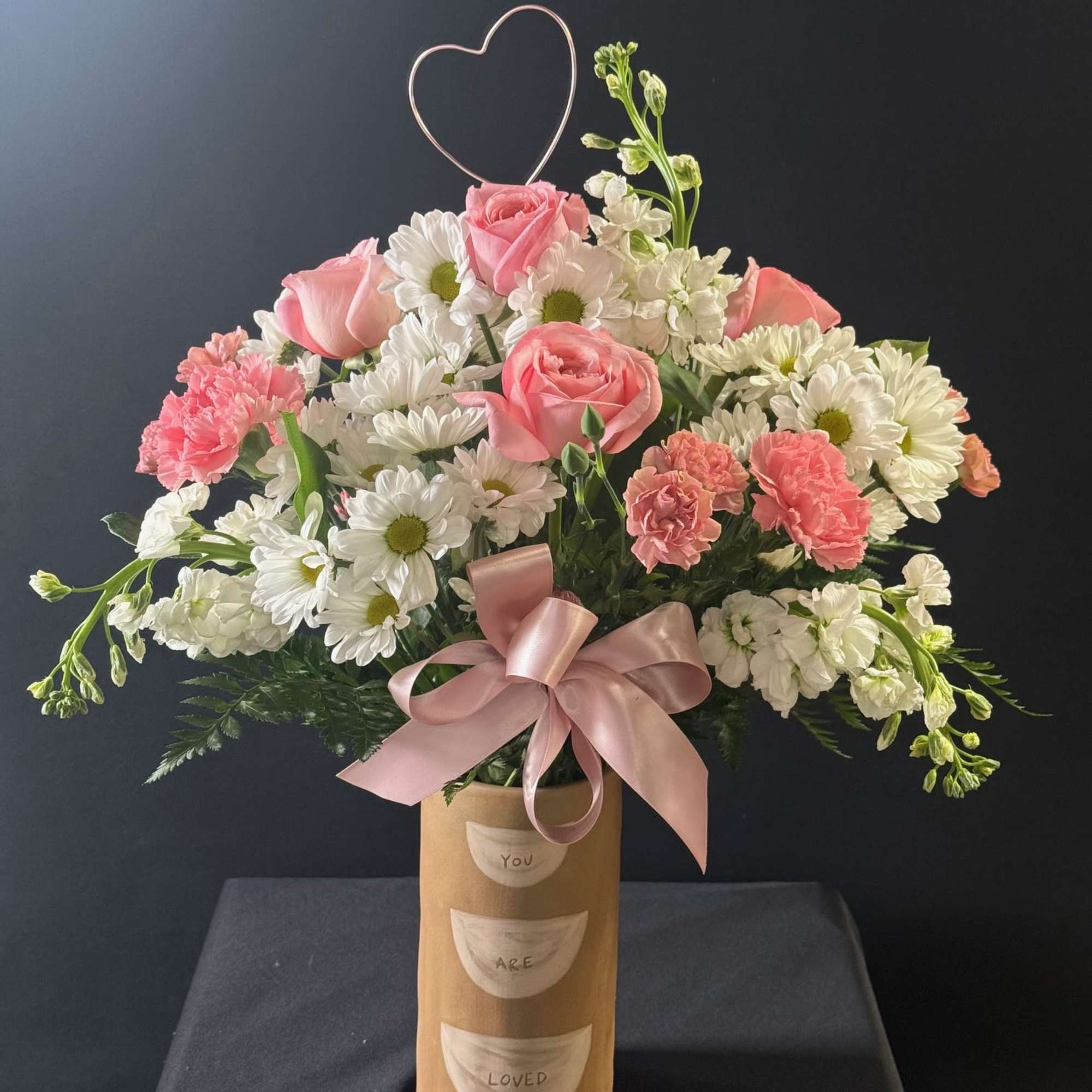 Pink roses and white daisies in a tall vase with a heart topper and ribbon