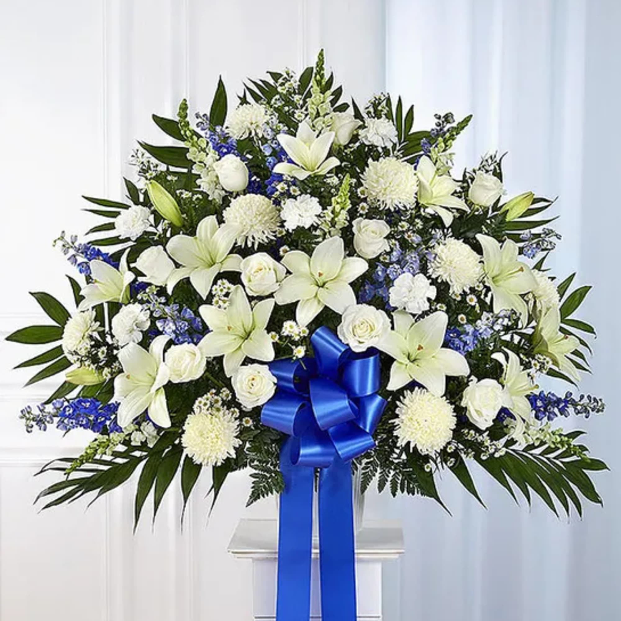 White lilies and roses arranged with blue flowers and a ribbon on a stand