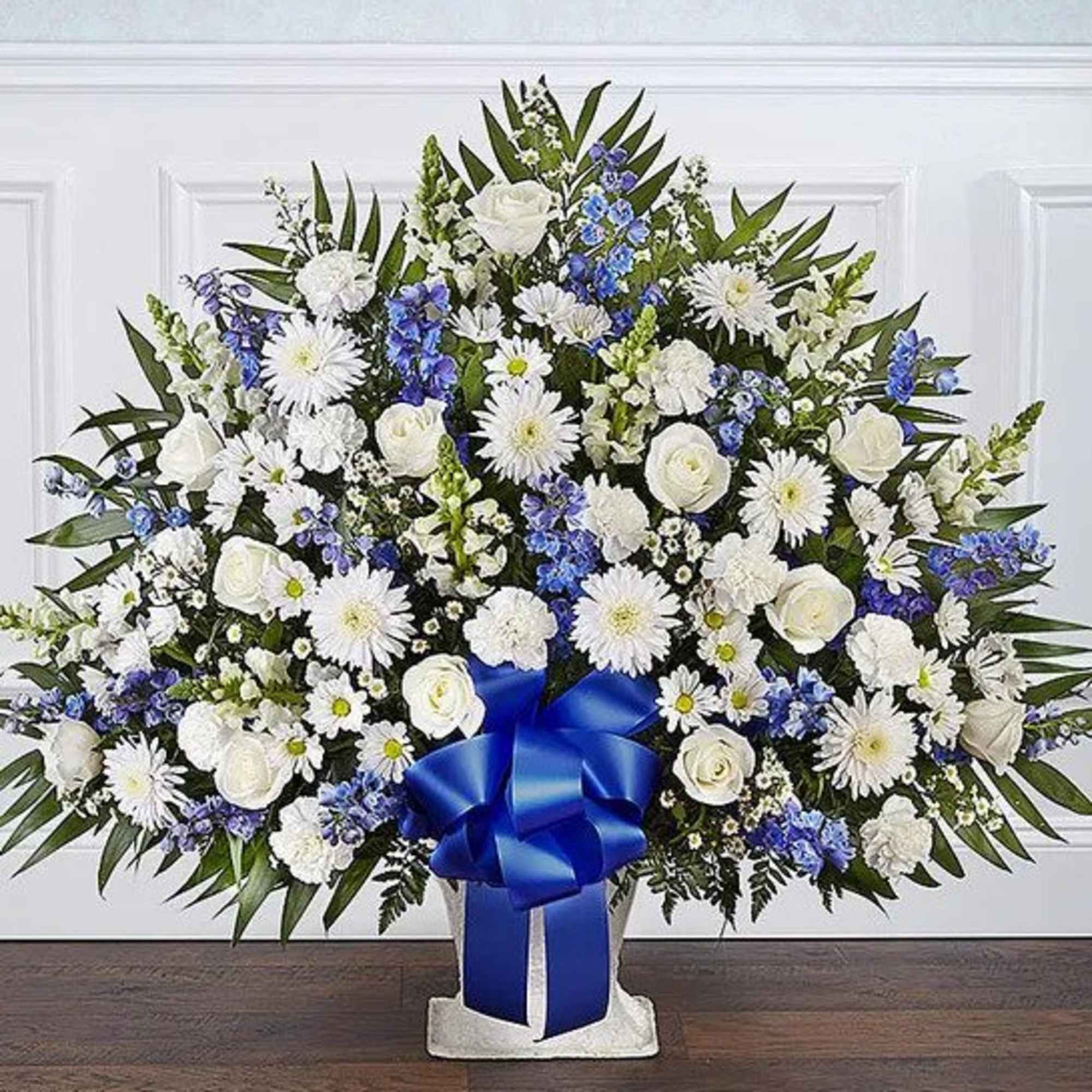 Large white and blue floral arrangement with roses, mums, and a bold blue ribbon bow in a white container