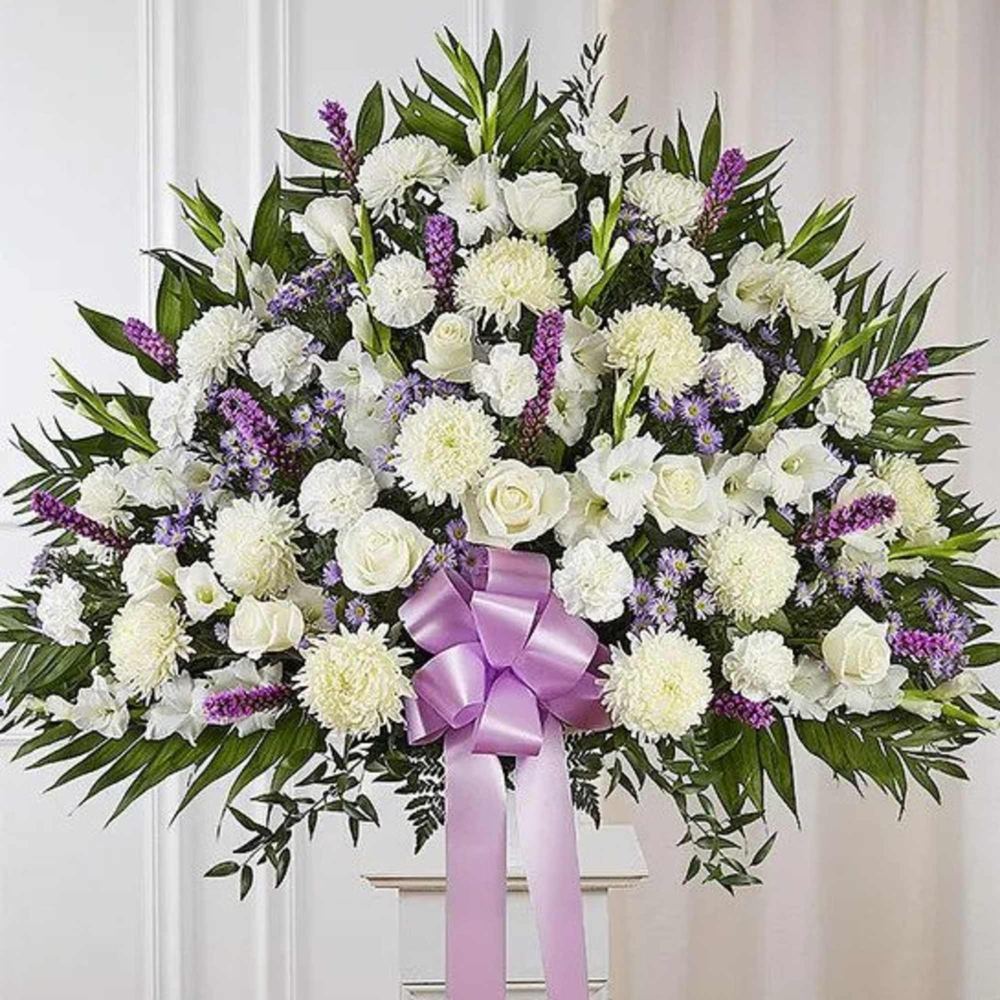 Large white and purple standing floral spray with roses, mums, and a lavender bow on a white pedestal