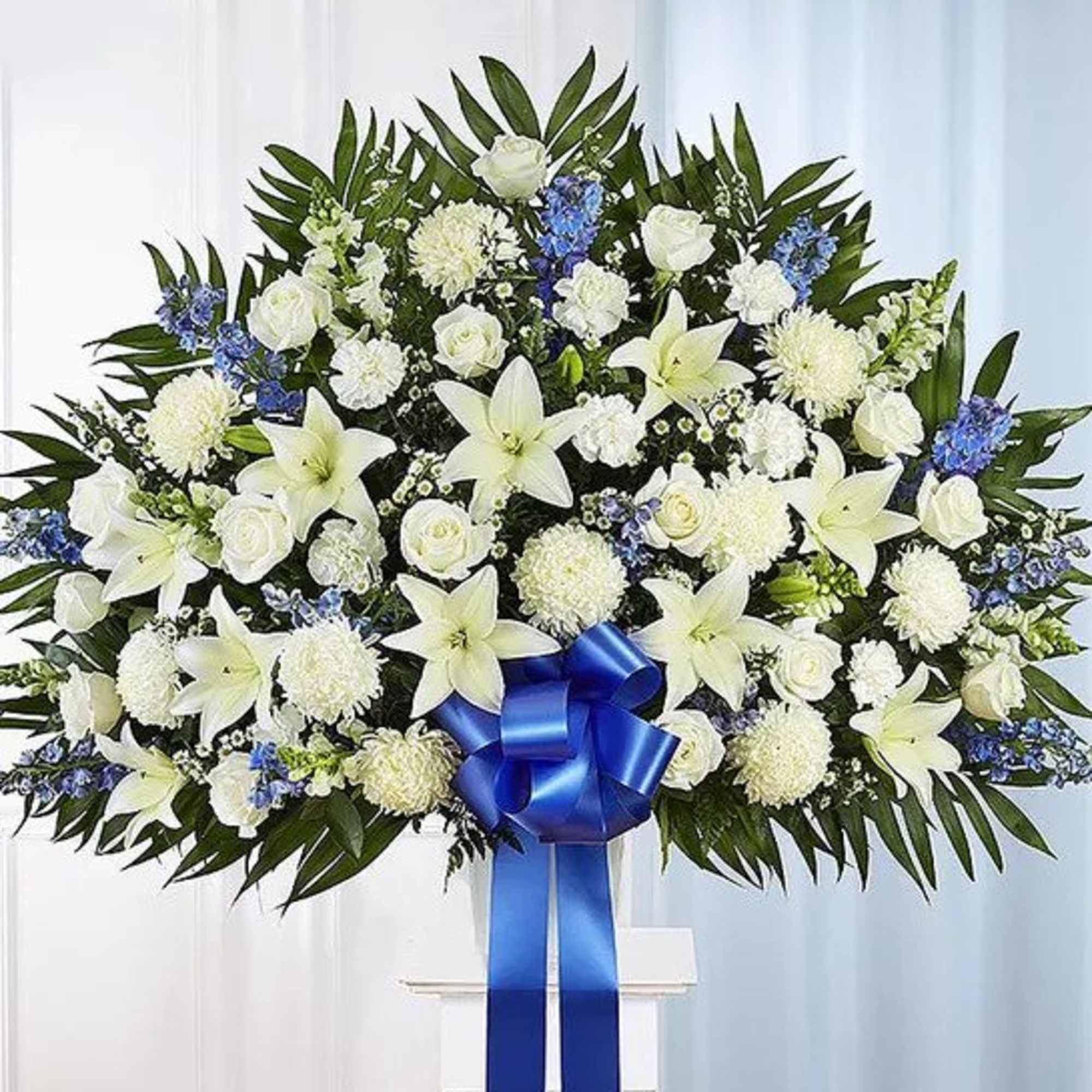 Large standing spray of white lilies, roses, mums, and blue flowers with a blue ribbon bow.