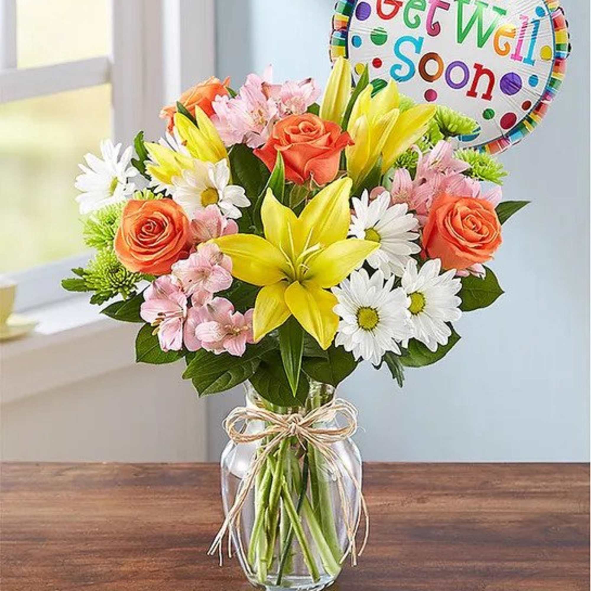 Mixed bouquet with yellow lilies, orange roses, white daisies and pink blooms in a clear vase with Get Well balloon