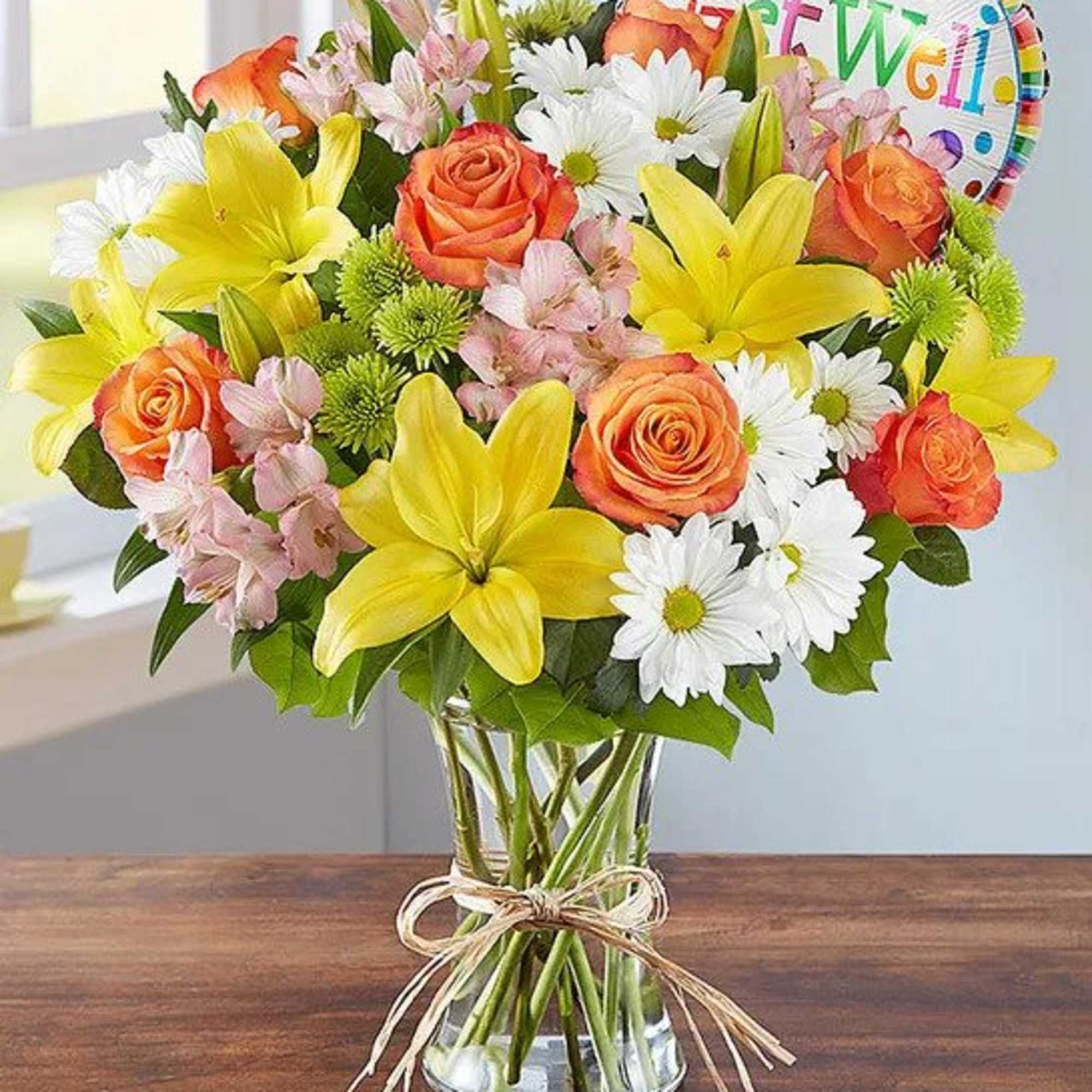 Mixed bouquet of yellow lilies, orange roses, white daisies, and pink flowers in a clear vase with a Get Well balloon