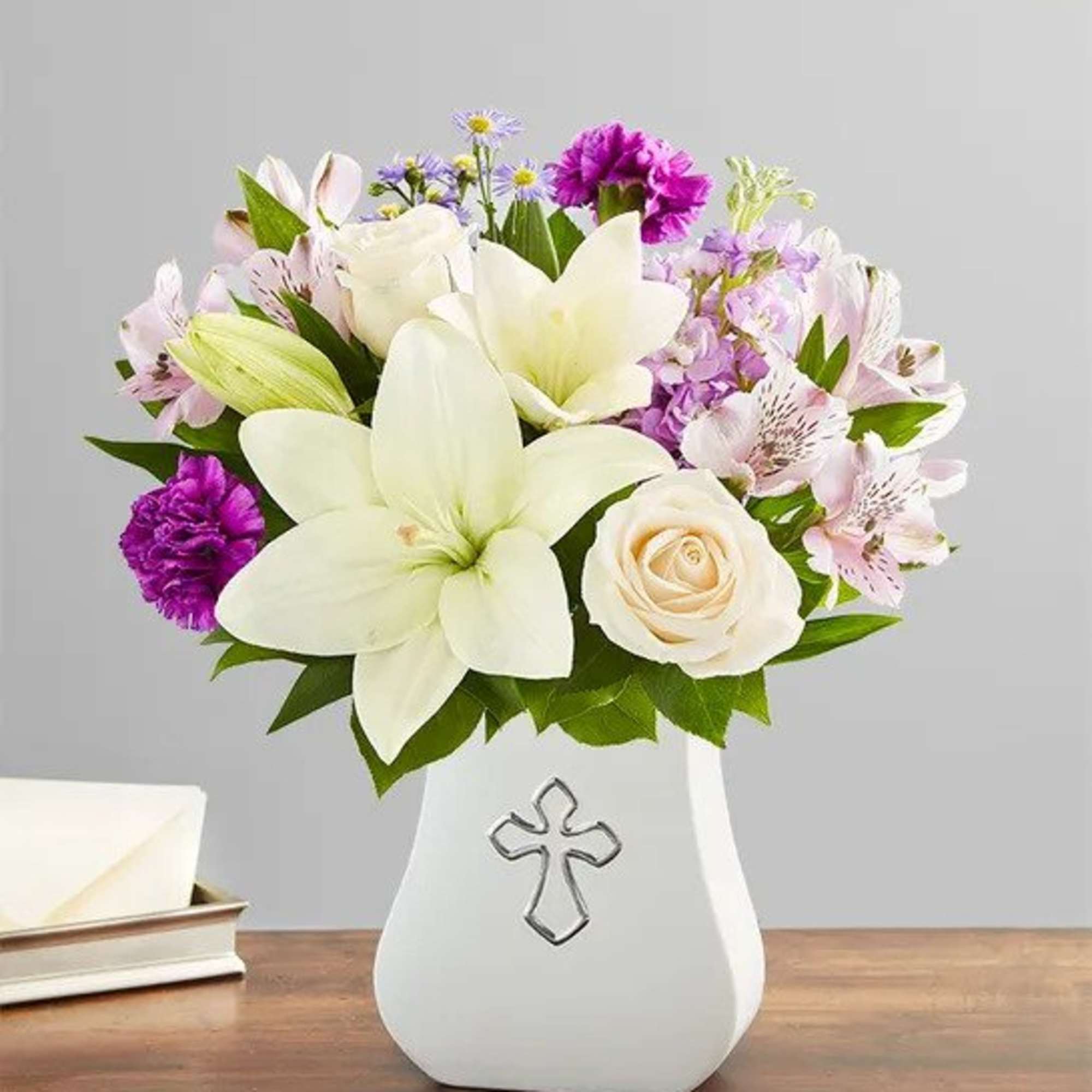 White lilies, roses, and purple flowers in a white vase with a silver cross design.