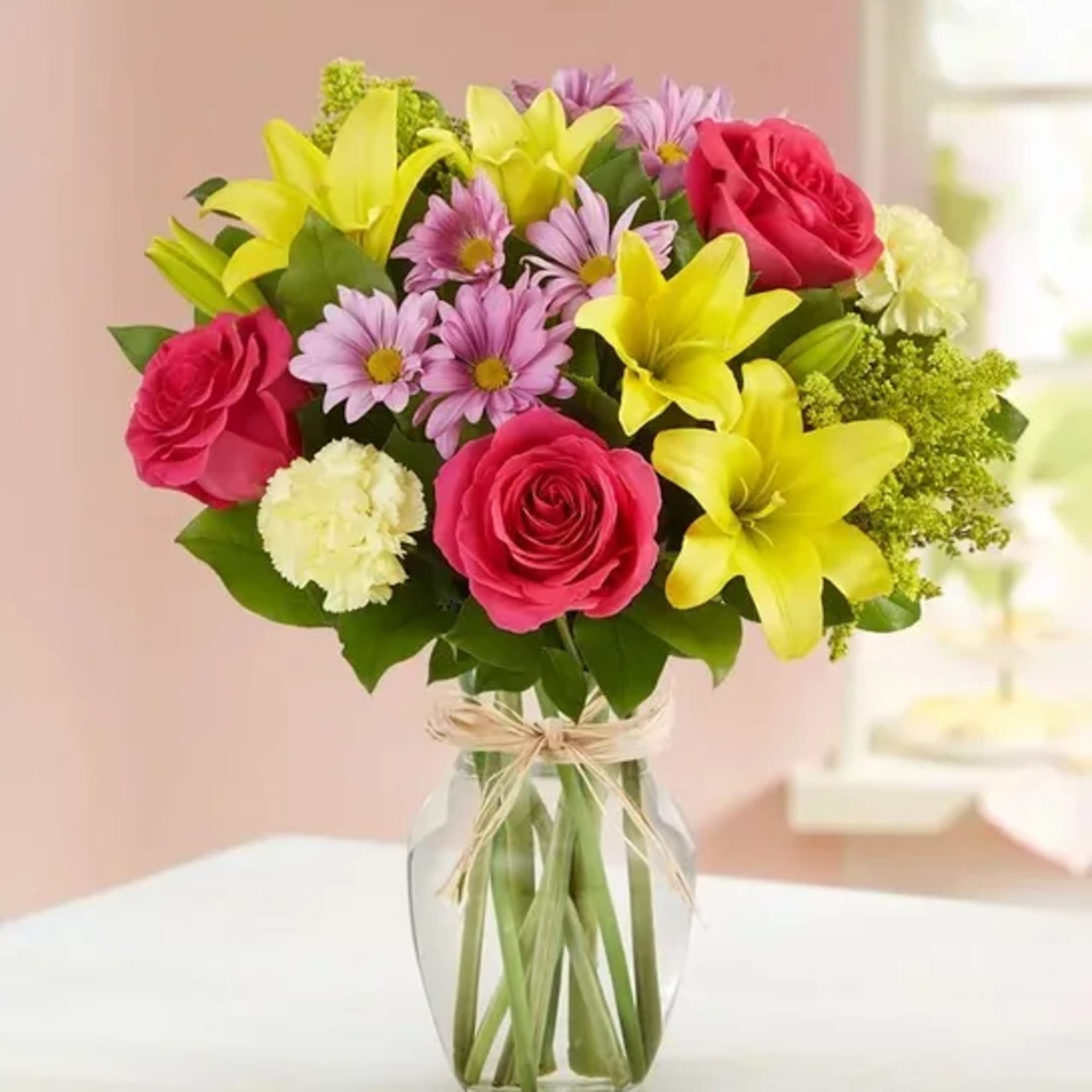 Mixed bouquet of yellow lilies, pink roses, lavender daisies, and pale carnations in a clear glass vase