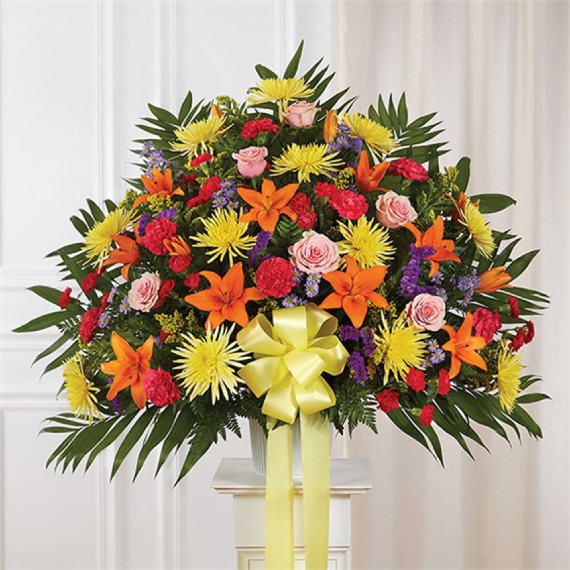 Large fan-shaped arrangement of bright mixed flowers with a yellow bow on a white pedestal.