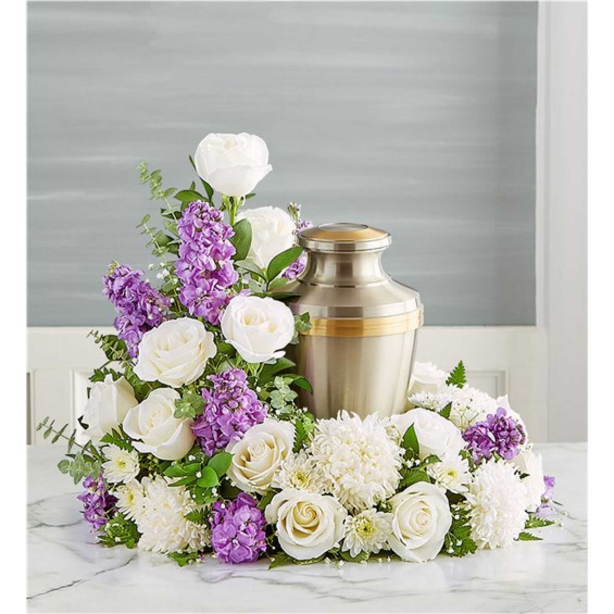 Cremation urn encircled by white roses, white mums, and purple flowers in a low arrangement.
