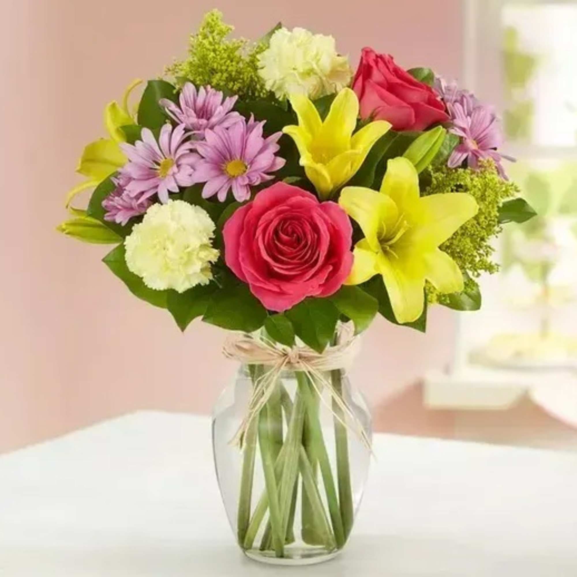 Mixed bouquet of pink roses, yellow lilies, purple daisies, and cream carnations in a clear glass vase