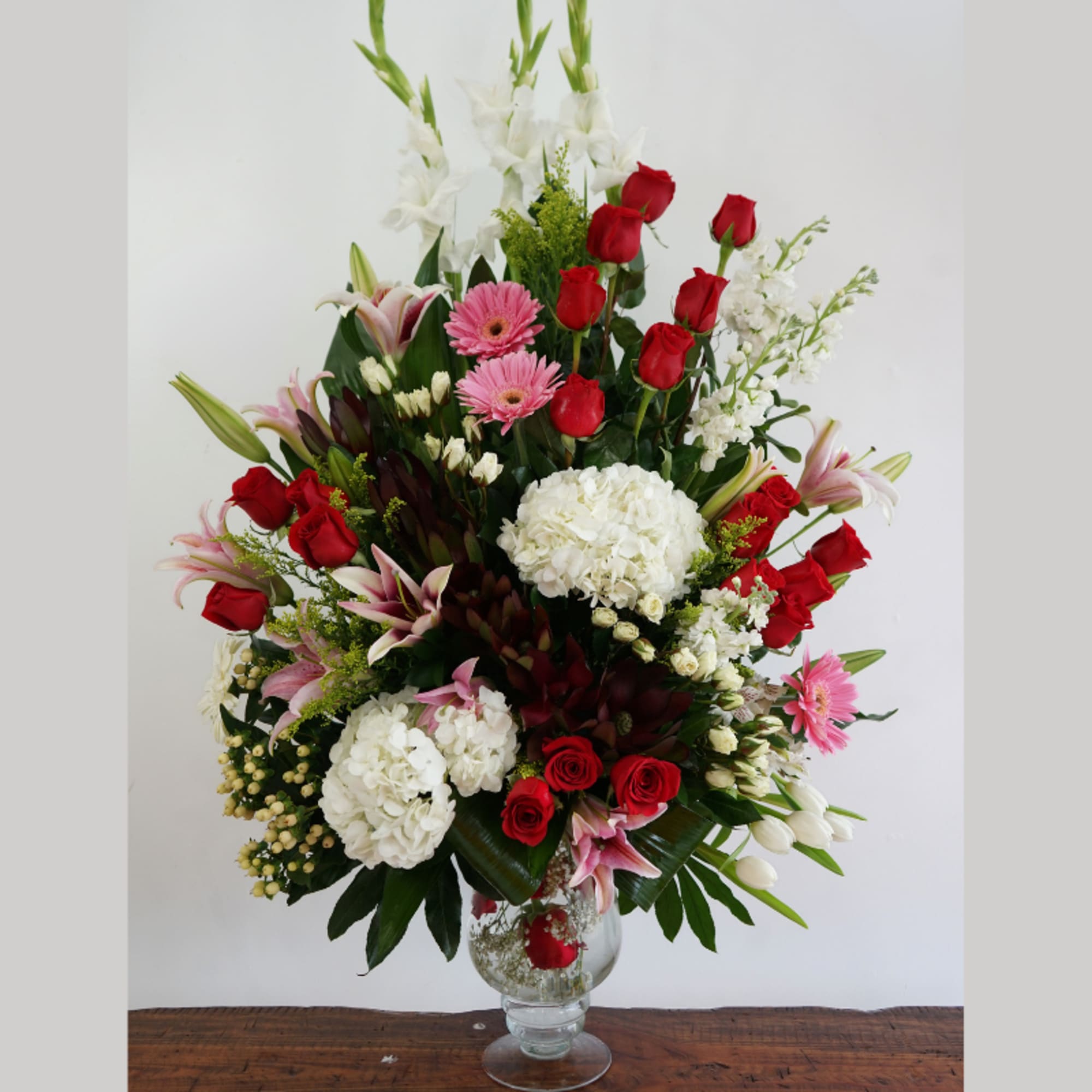 Tall mixed arrangement of red roses, white hydrangeas, pink lilies and gerberas in a clear glass vase