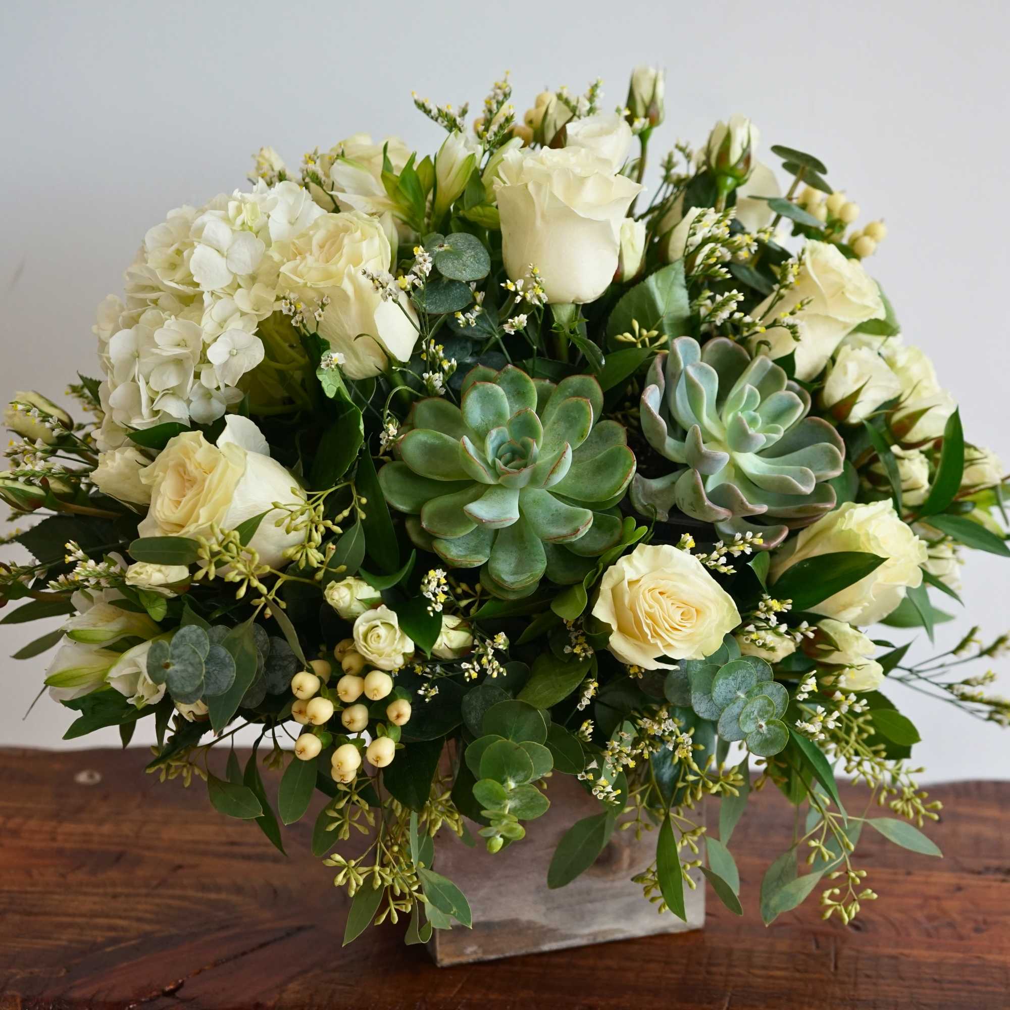 Compact arrangement of white roses, hydrangea, and succulents in a square vase.