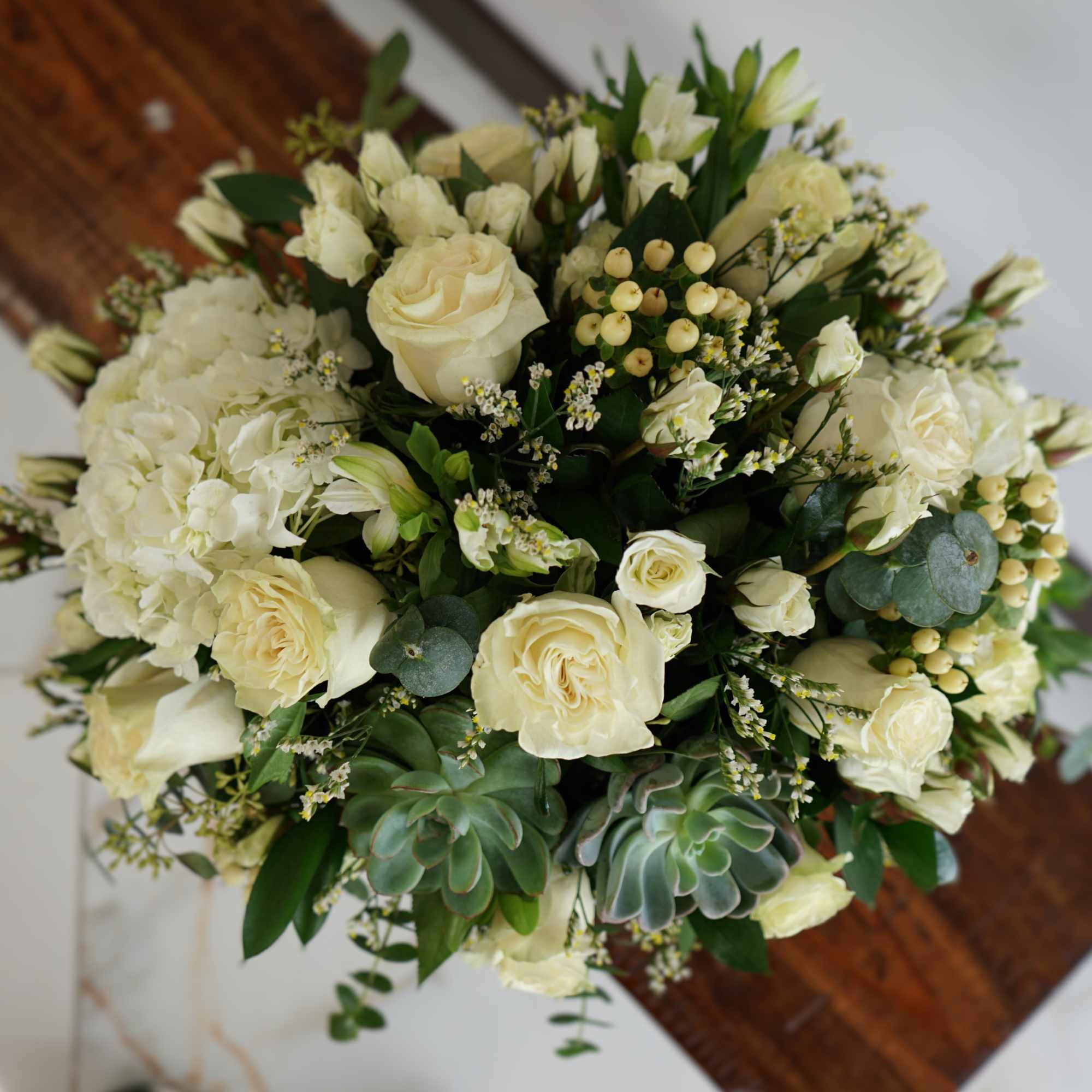 Round arrangement of cream roses, white hydrangeas, berries, and succulents viewed from above