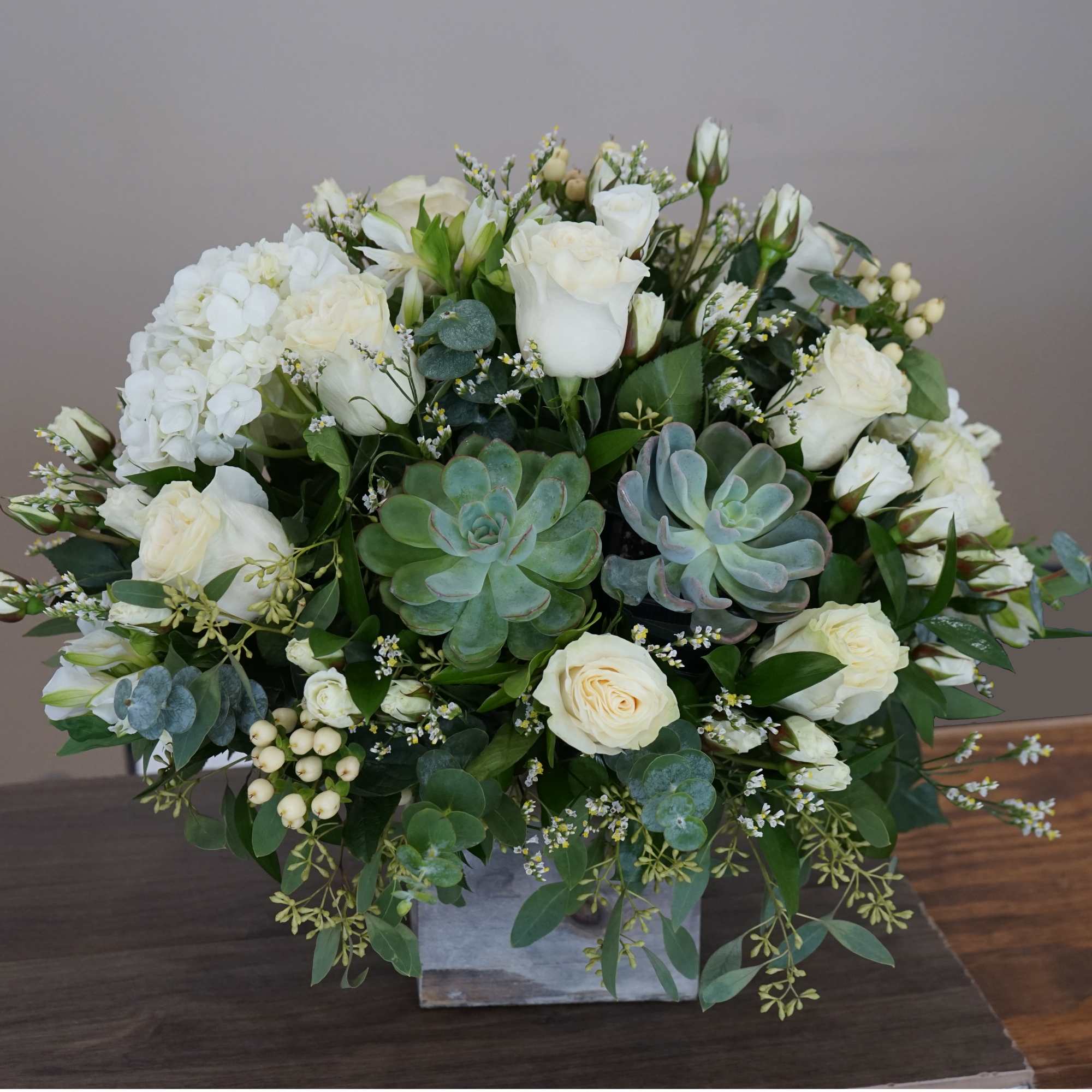 Low arrangement of white roses, hydrangea, and succulents in a square gray vase