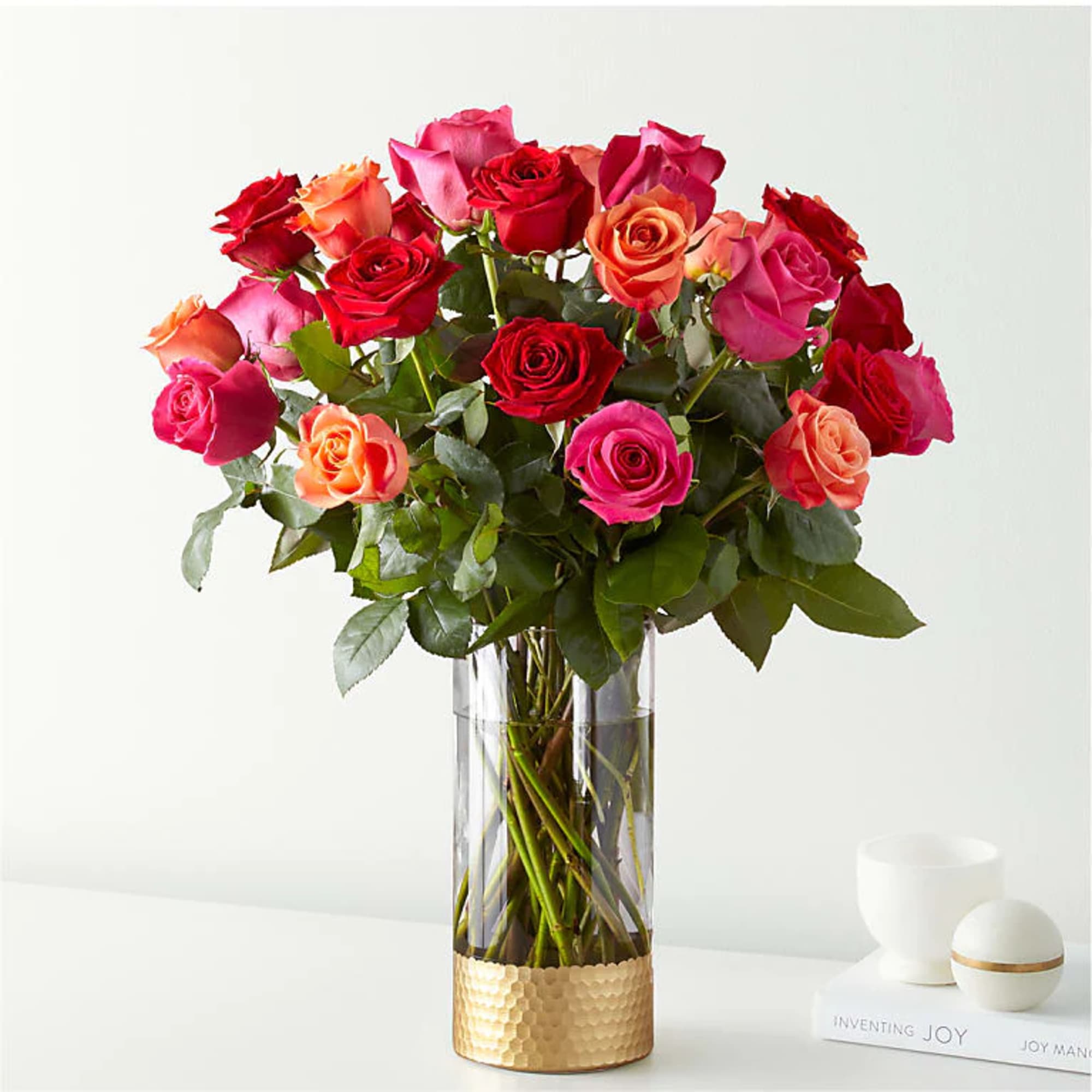 Tall bouquet of red, pink, and orange roses in a clear vase with a gold base