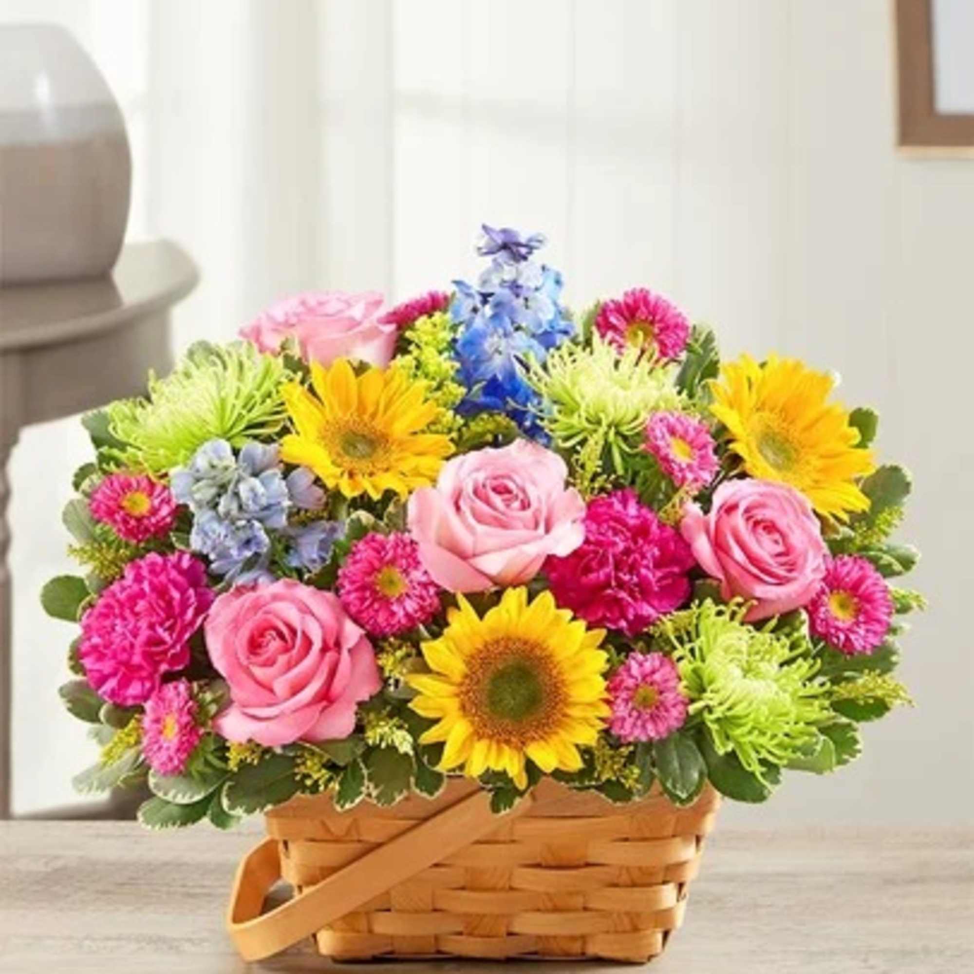 Bright basket arrangement with pink roses, yellow sunflowers, green mums, and blue accent flowers.