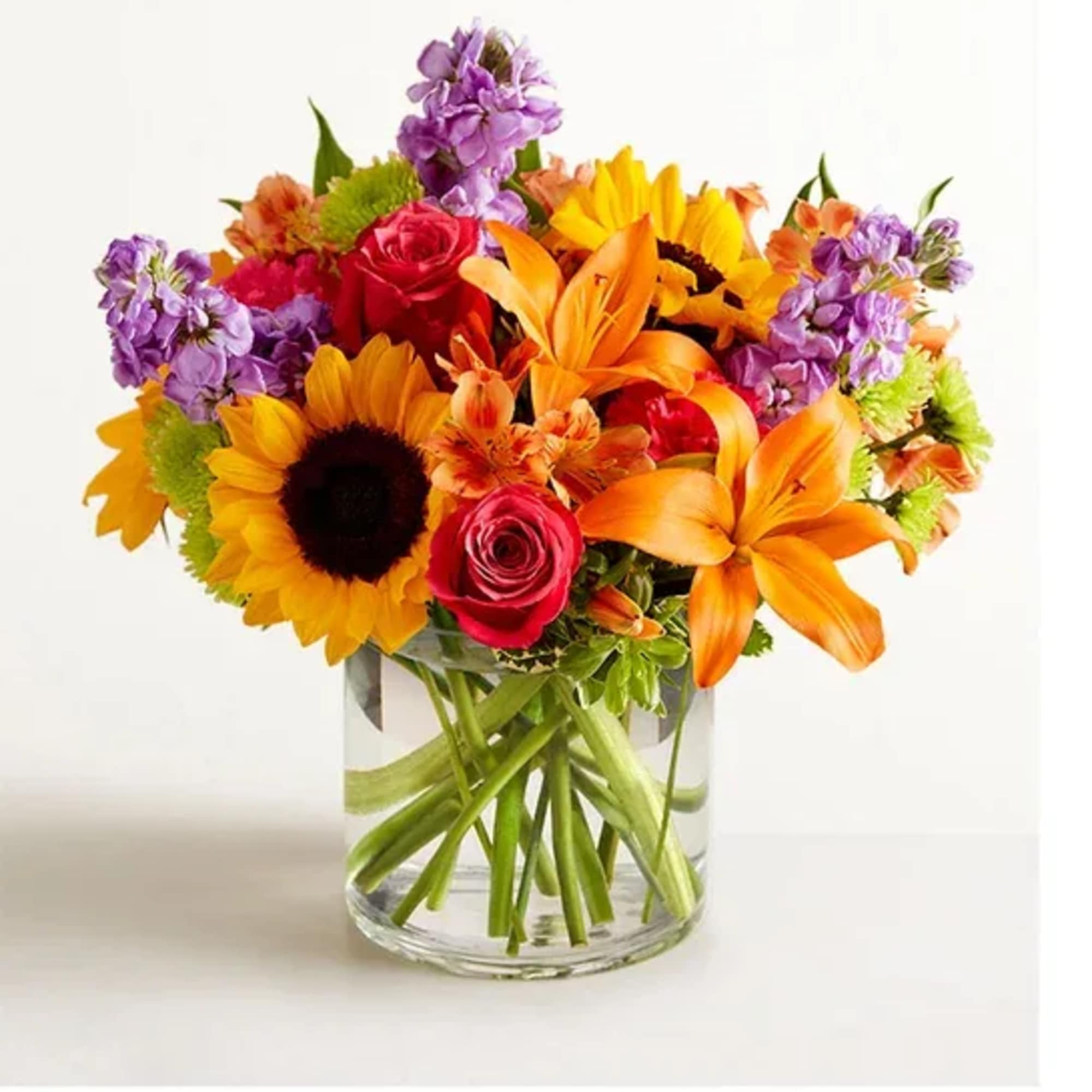 Bright mixed bouquet with sunflowers, orange lilies, roses, and purple flowers in a clear glass vase