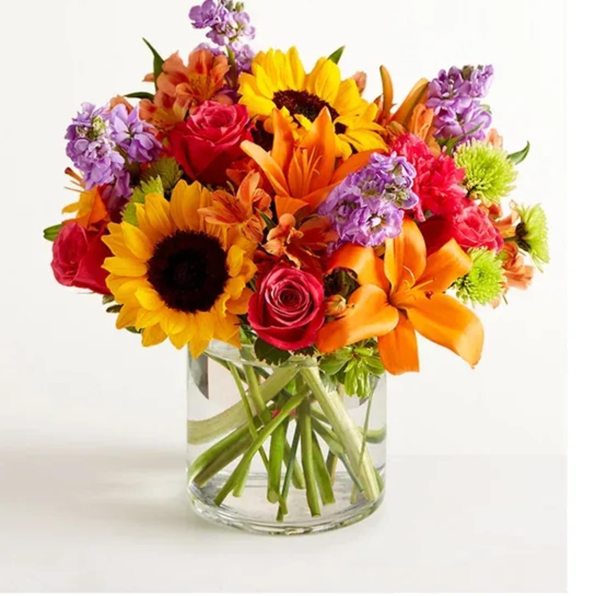 Bright mixed bouquet with sunflowers, roses, lilies, and purple blooms in a clear glass vase.