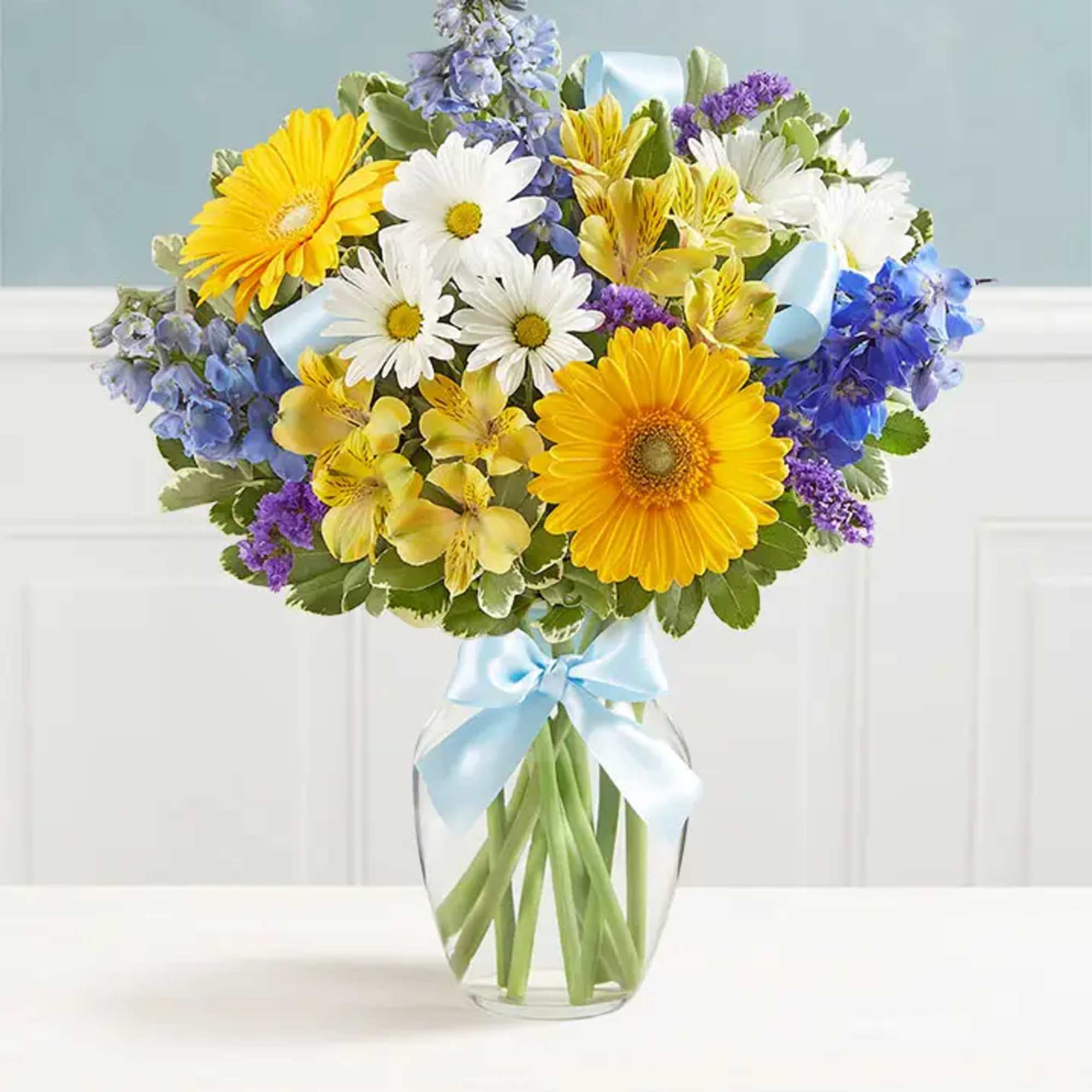 Bouquet of yellow, white, blue, and purple flowers in a glass vase with a blue ribbon