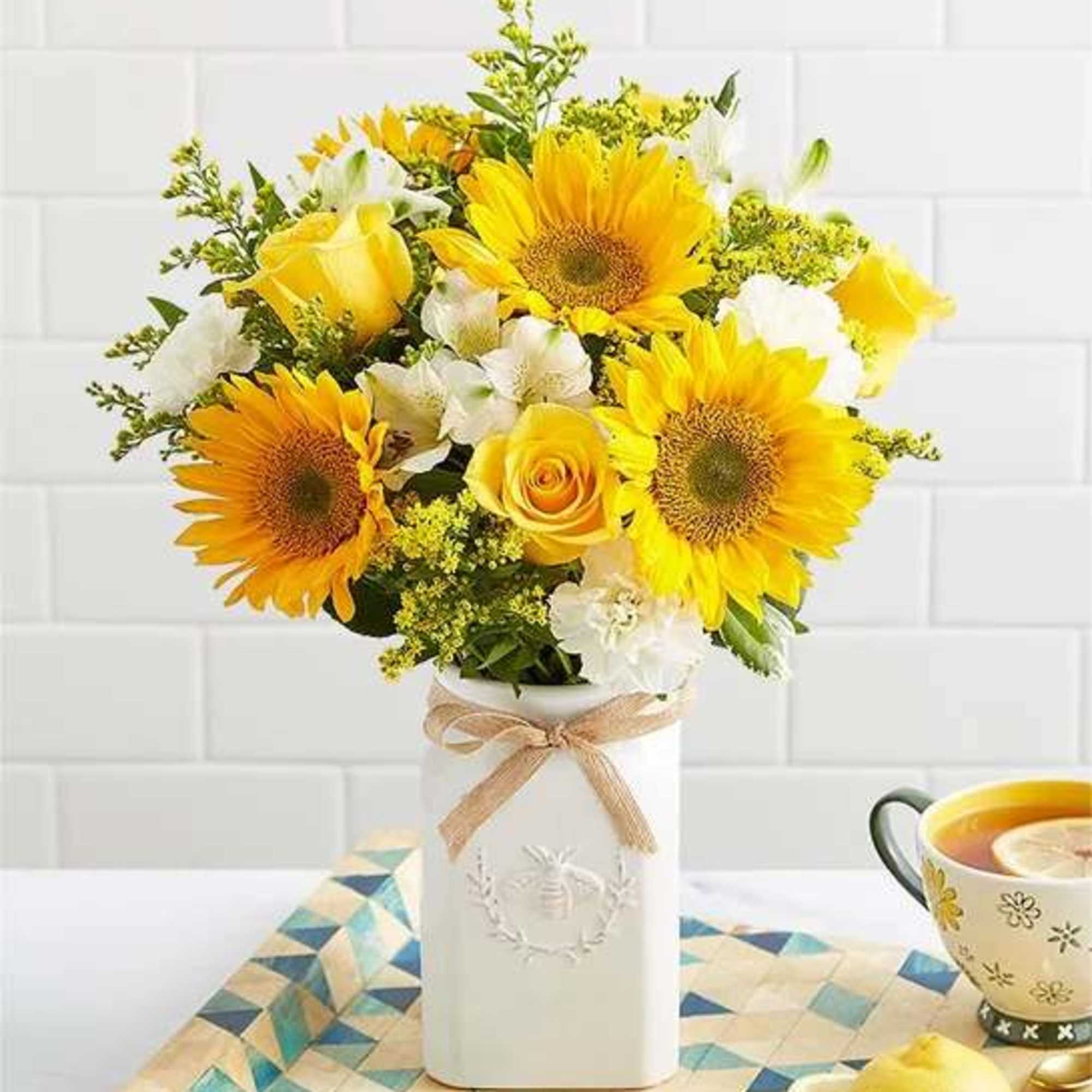 Yellow sunflowers and roses arranged in a white vase with a ribbon.