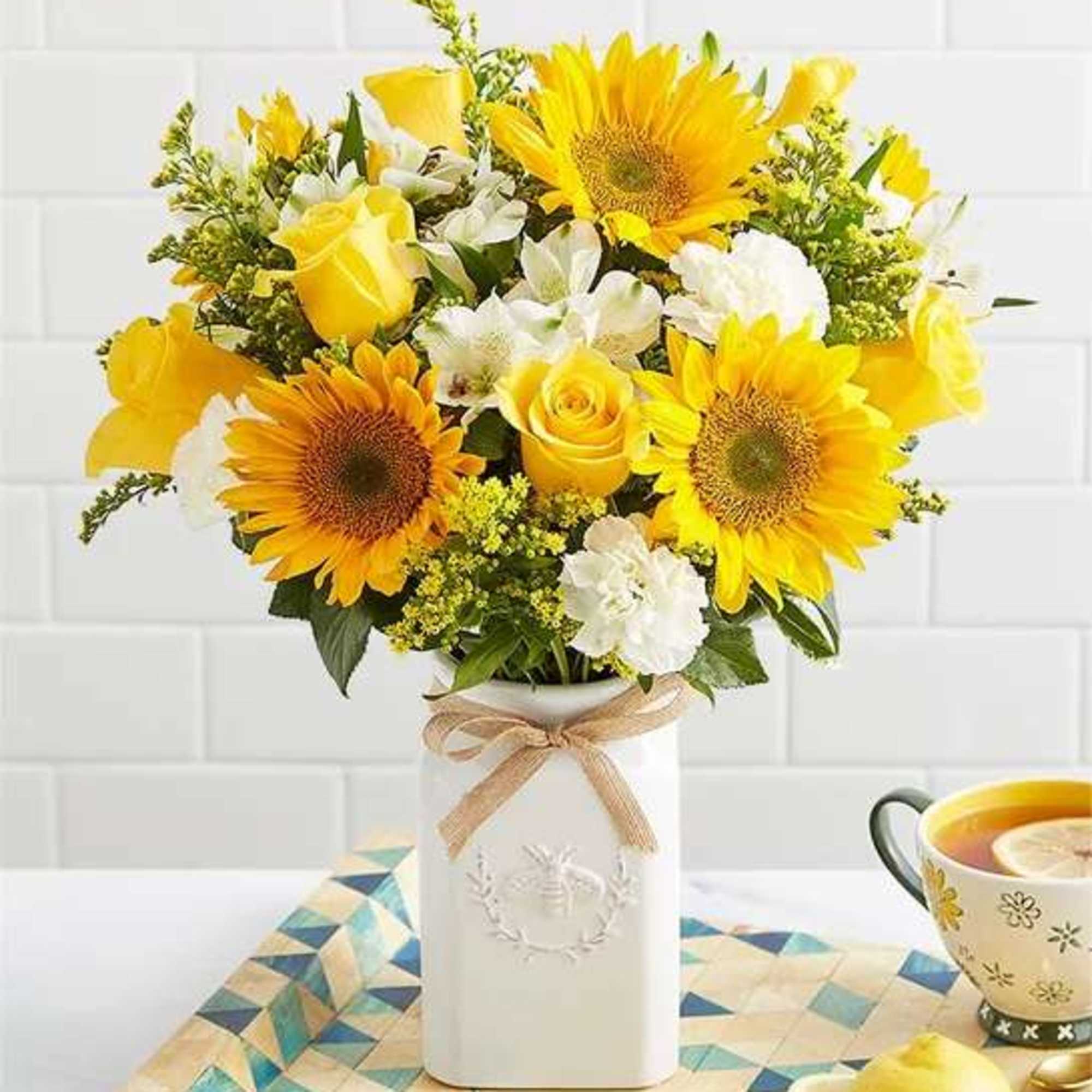 Yellow sunflowers and roses arranged in a white vase with a ribbon