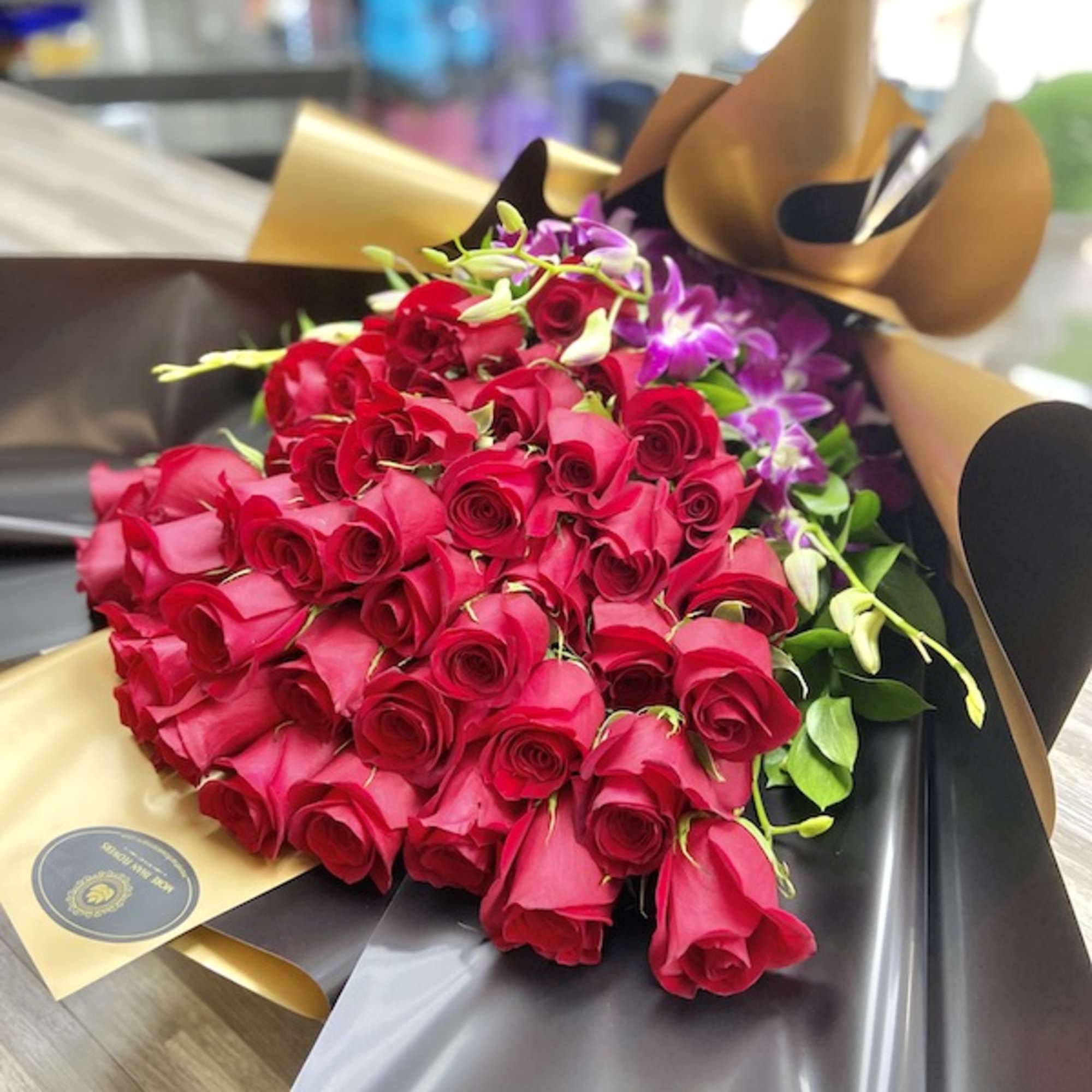 Large bouquet of red roses with purple orchids wrapped in black and gold paper on a table