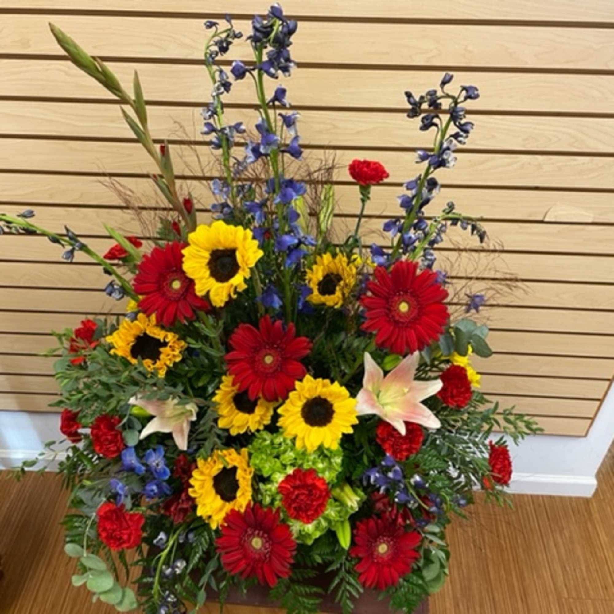 Tall mixed flower arrangement with sunflowers, red daisies, lilies, and blue delphinium