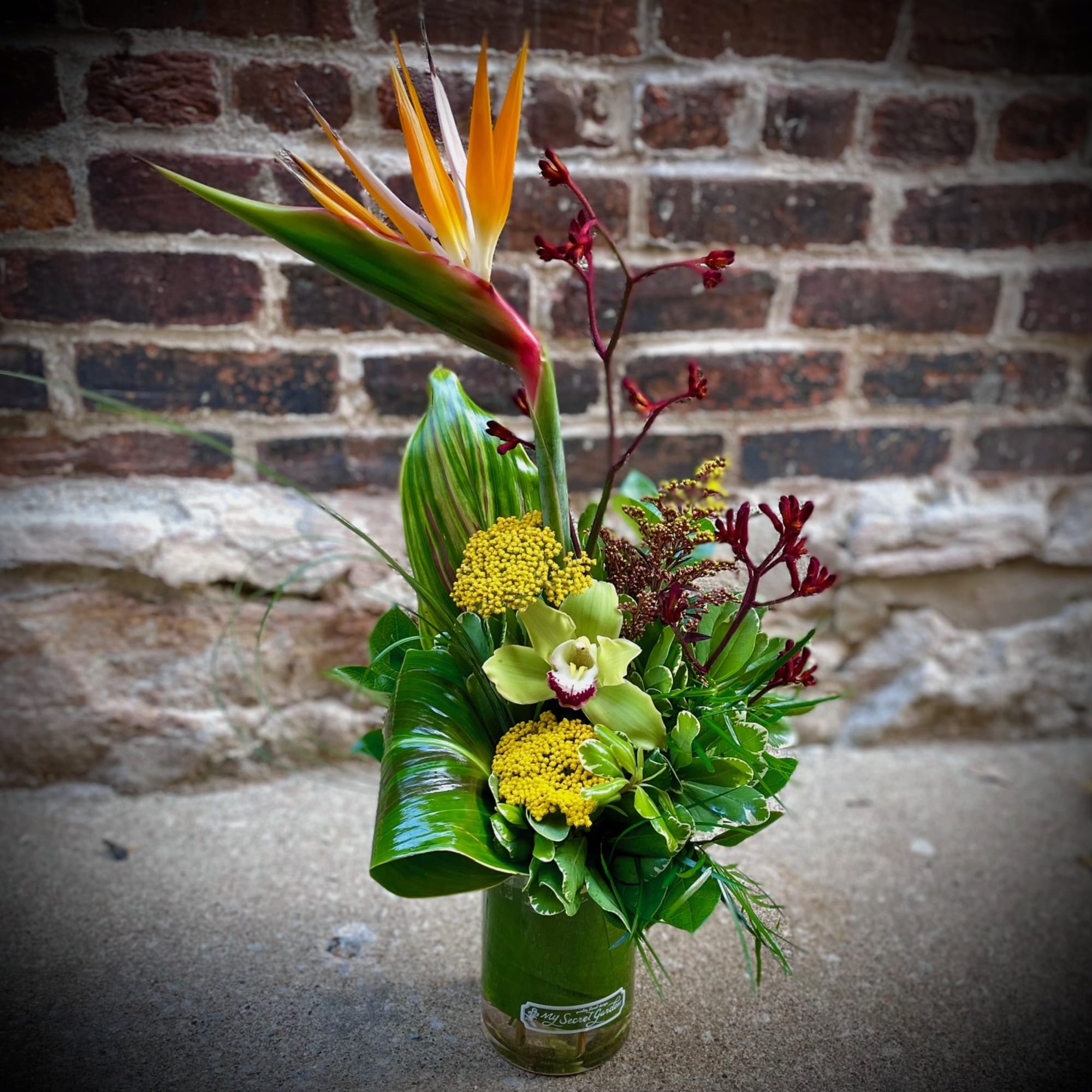 Tall tropical arrangement with bird of paradise, green orchid, and yellow blooms in a glass vase