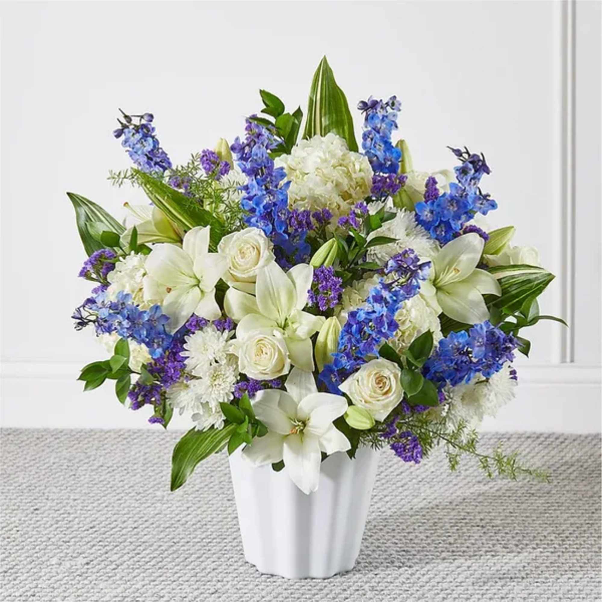 White lilies, roses, and blue flowers arranged in a white vase
