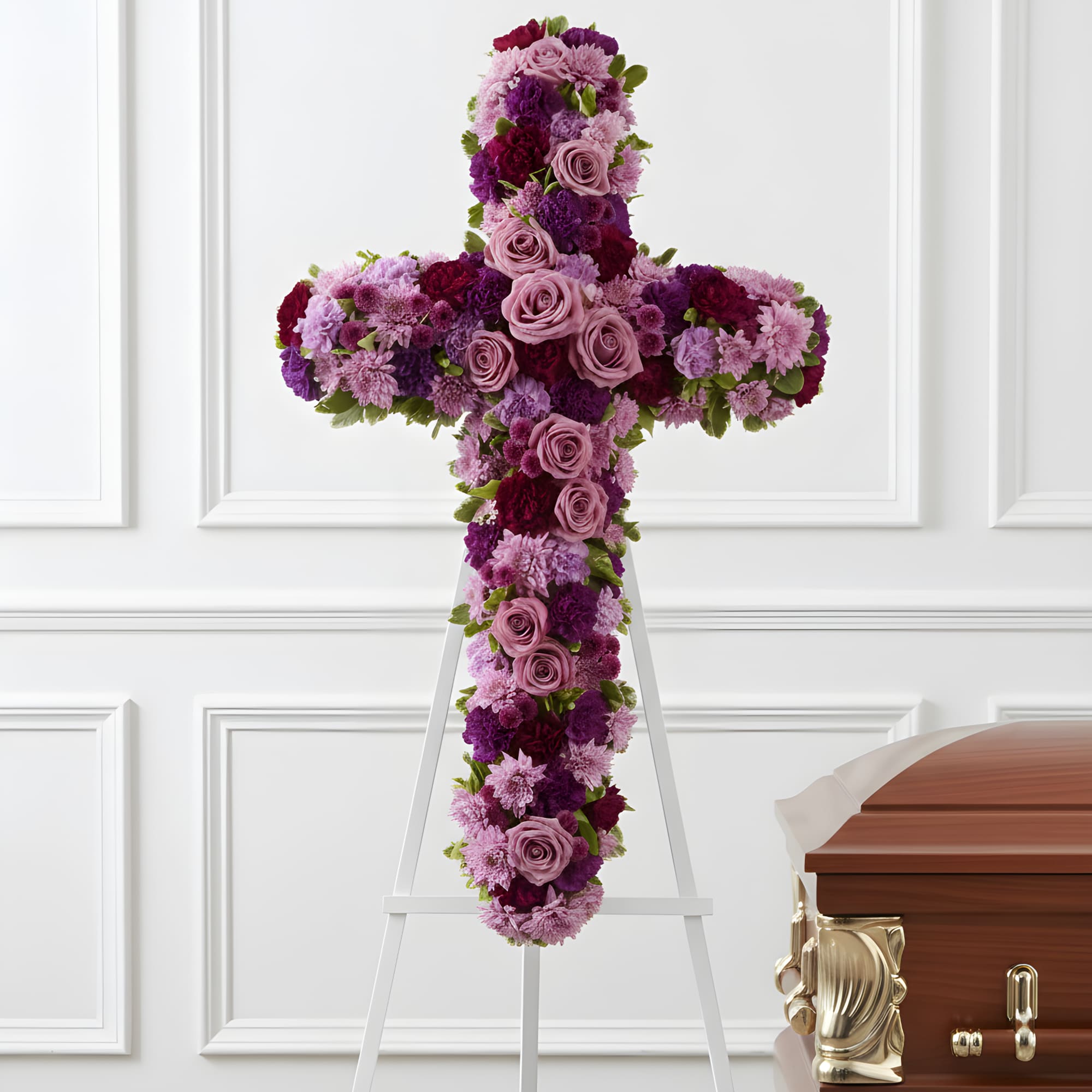 Purple and pink floral cross on a white easel beside a casket