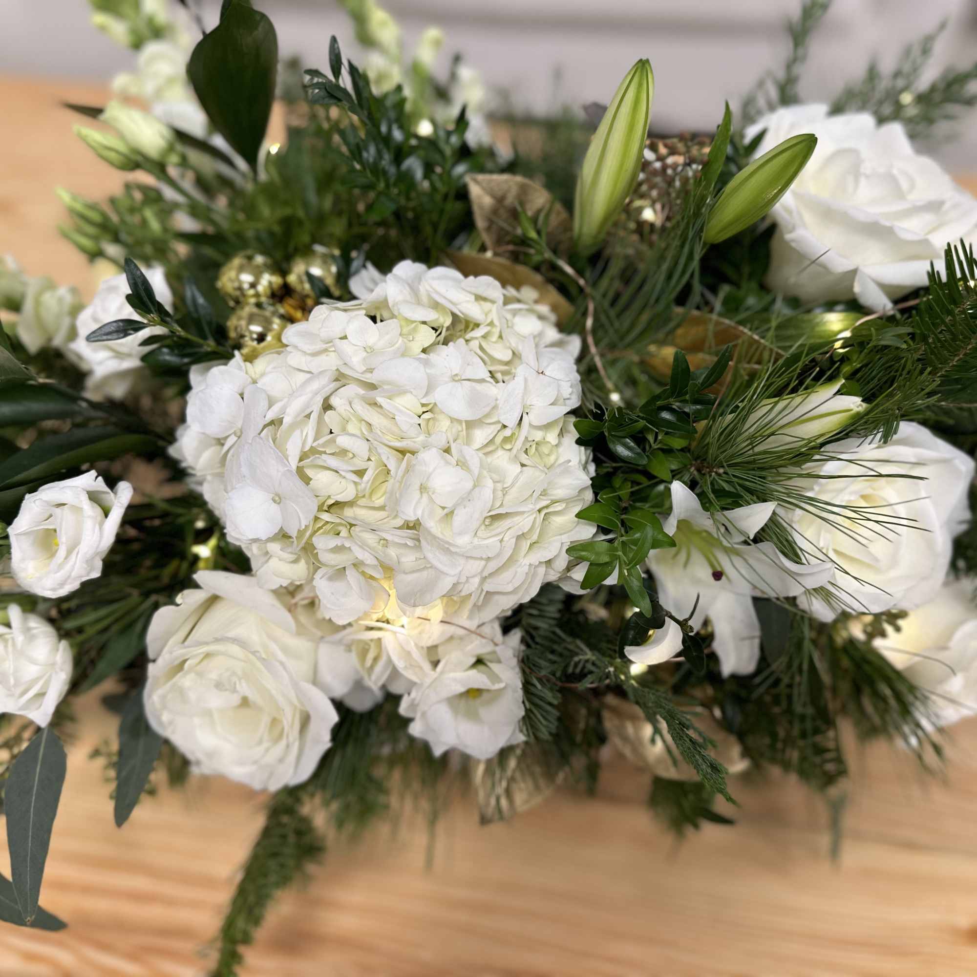 Low centerpiece of white hydrangea, roses, lilies, and lisianthus with evergreens and small lights.