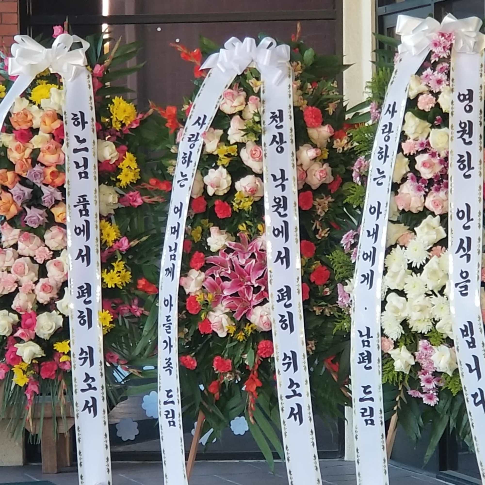 Three tall floral standing wreaths with white ribbon banners and mixed blooms